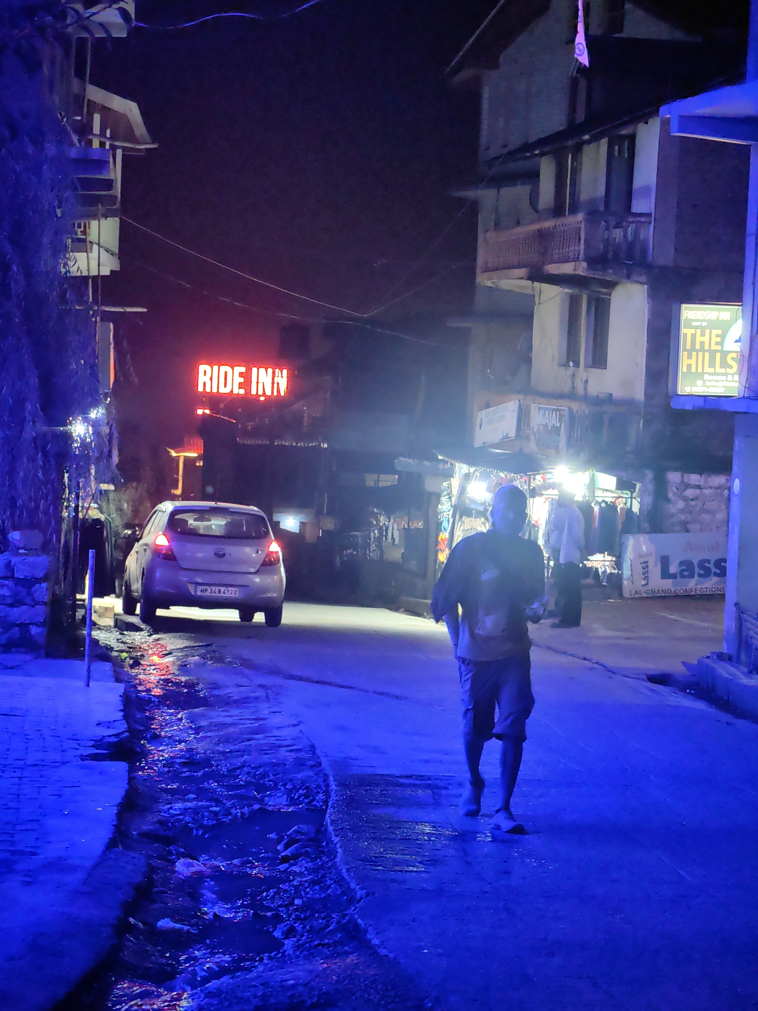 The image depicts a dimly lit street scene at night, likely in Barheta, India, illuminated by artificial light. In the foreground, a person walks away from the camera, wearing a t-shirt and shorts. To the left, a silver car is parked, its taillights illuminated. Above the car, a neon sign reads "RIDE INN" in red letters. Buildings line both sides of the street. On the right, a shop is visible with clothing racks displayed outside. A sign for "Lassi" is prominent on the right side of the street. The street appears wet, with water reflecting the colored lights, suggesting recent rain or a flowing gutter. The overall atmosphere is that of a developing or less affluent urban area at night.