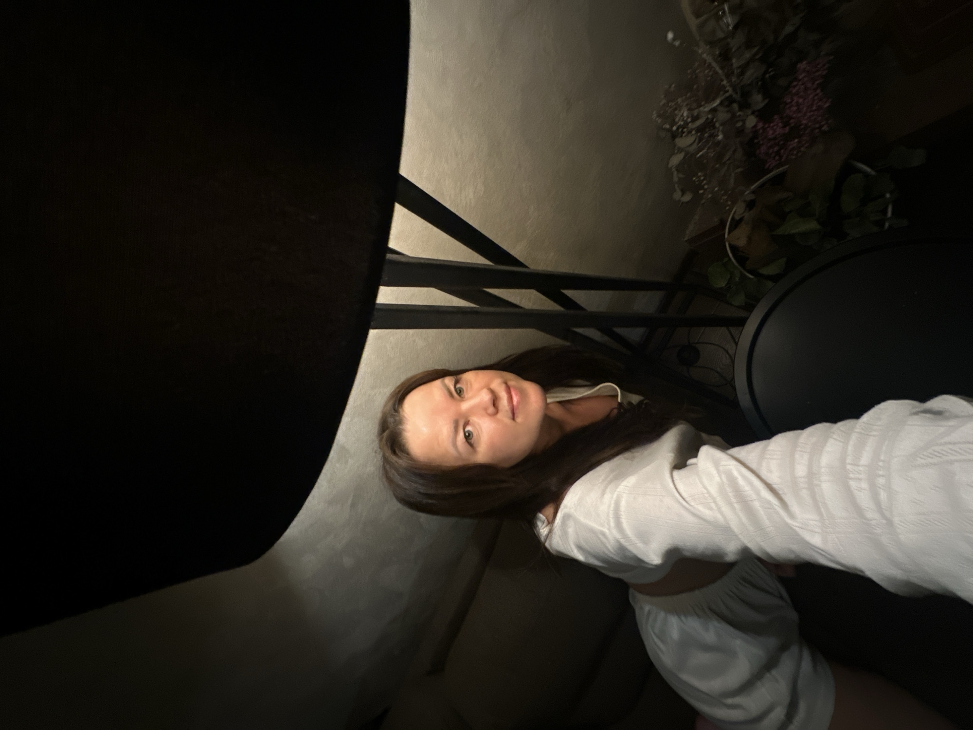 An indoor portrait taken in Chaikovsky, Russia, features a young woman looking directly at the camera with a calm, pleasant expression and a slight smile. She has long, dark hair and is wearing a light-colored, possibly white, long-sleeved top and matching shorts.
The setting appears to be a dimly lit room, suggesting it is evening or night. The woman is positioned on a dark-colored couch or seating. A large, dark object, likely a lampshade, is prominent in the foreground to the upper left, casting a soft glow on the wall behind the woman. This wall is light-colored and textured.
In the background, to the woman's right, there's a decorative arrangement of what looks like dried flowers or foliage, showing hints of purple and white, placed on a dark, circular surface, possibly a table. Metal structural elements, perhaps part of shelving or furniture, are also visible. The overall mood of the image is relaxed and candid. No visible text is present.