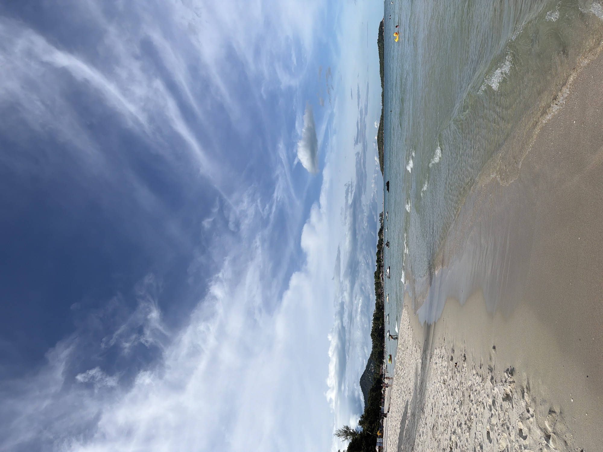 This is a photograph of a tropical beach on a sunny day. The foreground is dominated by a sandy beach with footprints. The ocean water is clear and calm, with small waves gently lapping at the shore. In the middle ground, several people are visible on the beach and in the water. Some are walking along the shoreline, while others are swimming or using inflatable toys. Several jet skis are parked or in use in the water. In the distance, lush green hills and a line of trees form a backdrop to the beach. The sky is a vibrant blue, dotted with wispy white clouds. The overall scene depicts a lively and picturesque beach resort. This appears to be a scene from Baan Chaweng, Thailand, a popular tourist destination known for its beautiful beaches and clear waters.
