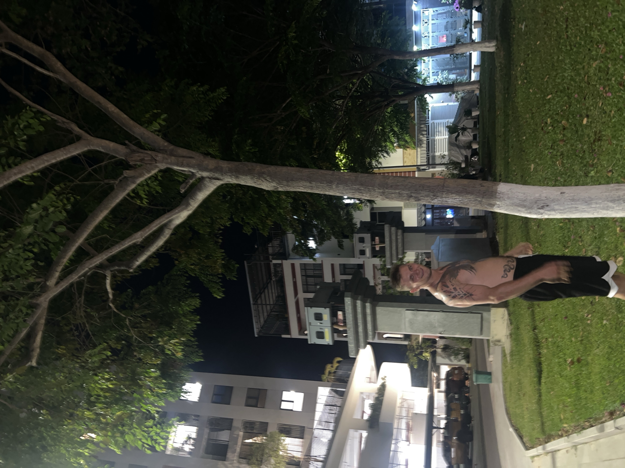 This nighttime photograph captures a shirtless man posing in a landscaped area, likely in the Nam Natural Heritage Area Trang, Vietnam, as indicated by the context. The man stands on a vibrant green lawn, smiling directly at the camera. He wears dark shorts with a white trim and displays prominent tattoos on his left shoulder/upper arm and a snake-like design on his torso.
In the foreground, a large tree with light-colored, branching limbs and sparse green leaves dominates the left side of the frame. Other trees, some with white-painted trunks, are visible in the mid-ground and background, contributing to a park-like atmosphere. Beyond the trees and grassy areas, several modern, multi-story buildings with illuminated windows and balconies create the backdrop, suggesting a residential or urban environment. A paved path or road runs along the mid-ground, separating the lawn from the buildings.
The scene is lit by artificial lights from the buildings and possibly streetlights, casting a warm glow on certain areas and contrasting with the dark night sky. The overall impression is one of a relaxed evening in a well-maintained outdoor space within an urban setting. No distinct legible text is visible in the image.