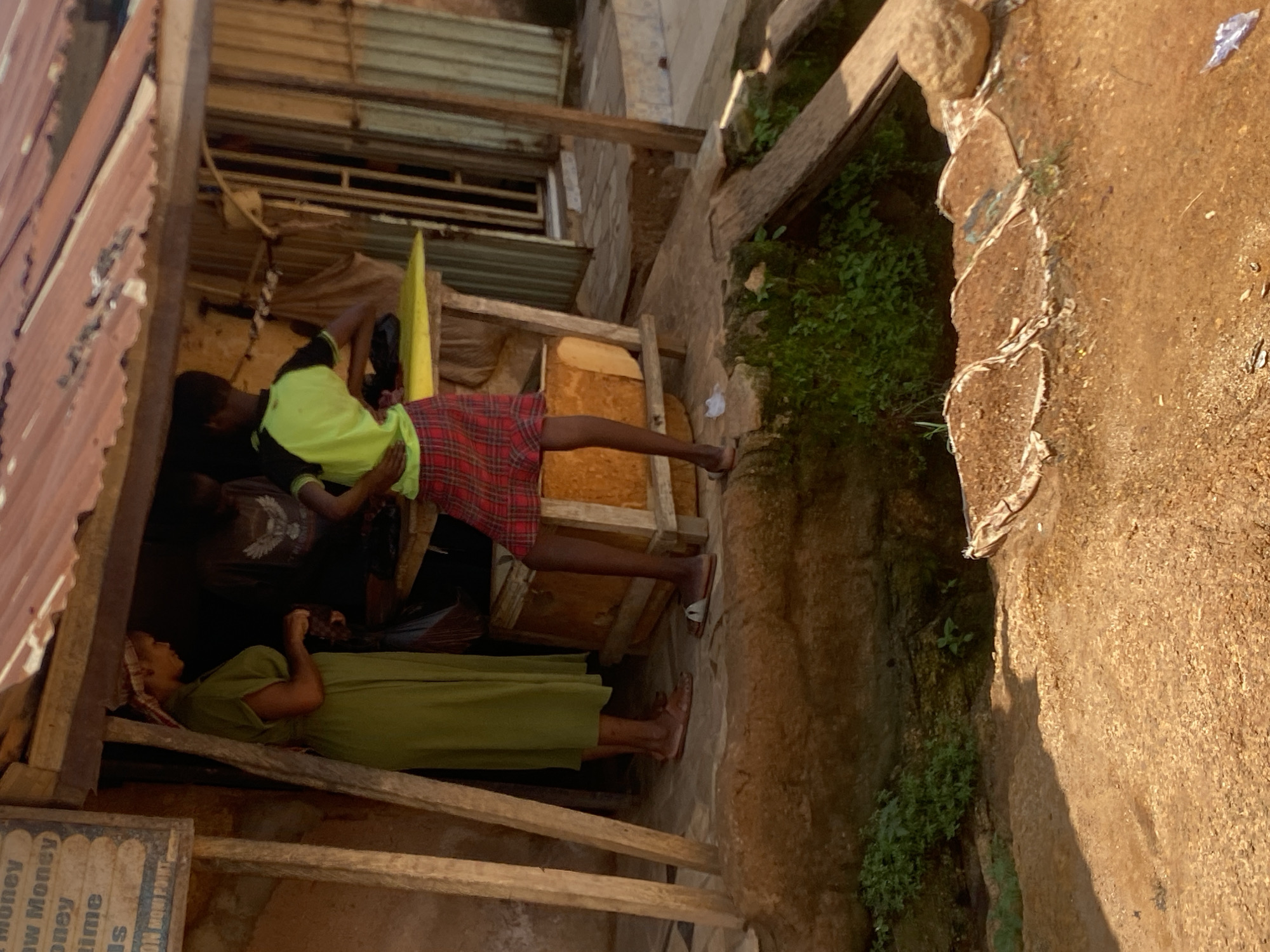 The image depicts an outdoor scene in what appears to be a humble dwelling or market stall, possibly in Jos, Nigeria, given the provided context. The perspective is slightly tilted, making it feel dynamic.
In the foreground, a person in a vibrant green dress is lying down, resting on a pillow. Their head is turned away from the viewer, and they appear to be relaxed or asleep. Behind them, another person is seated, wearing a bright green t-shirt and a red and black plaid skirt. This individual is leaning against some wooden structures and seems to be engaged in a task, possibly sorting or handling something. A third person, partially visible and wearing a dark shirt, is also present.
The setting is characterized by rustic wooden structures, corrugated metal roofing, and what looks like a rough, earthy wall or natural rock formation to the right. Green foliage is visible growing in crevices of the wall, suggesting an outdoor environment that might be prone to some overgrowth. The lighting indicates it is daytime, with sunlight casting shadows and illuminating the textures of the materials.
There is a sign with some visible text on the lower left, appearing to be written in white on a darker background. The visible words include "Money", "Now Money", "oney", and "is". It's possible this is a sign for a money lending or exchange service.
The overall impression is one of everyday life in a simple, possibly crowded, environment, with people taking a moment to rest or attend to their activities. The scene feels authentic and unposed.