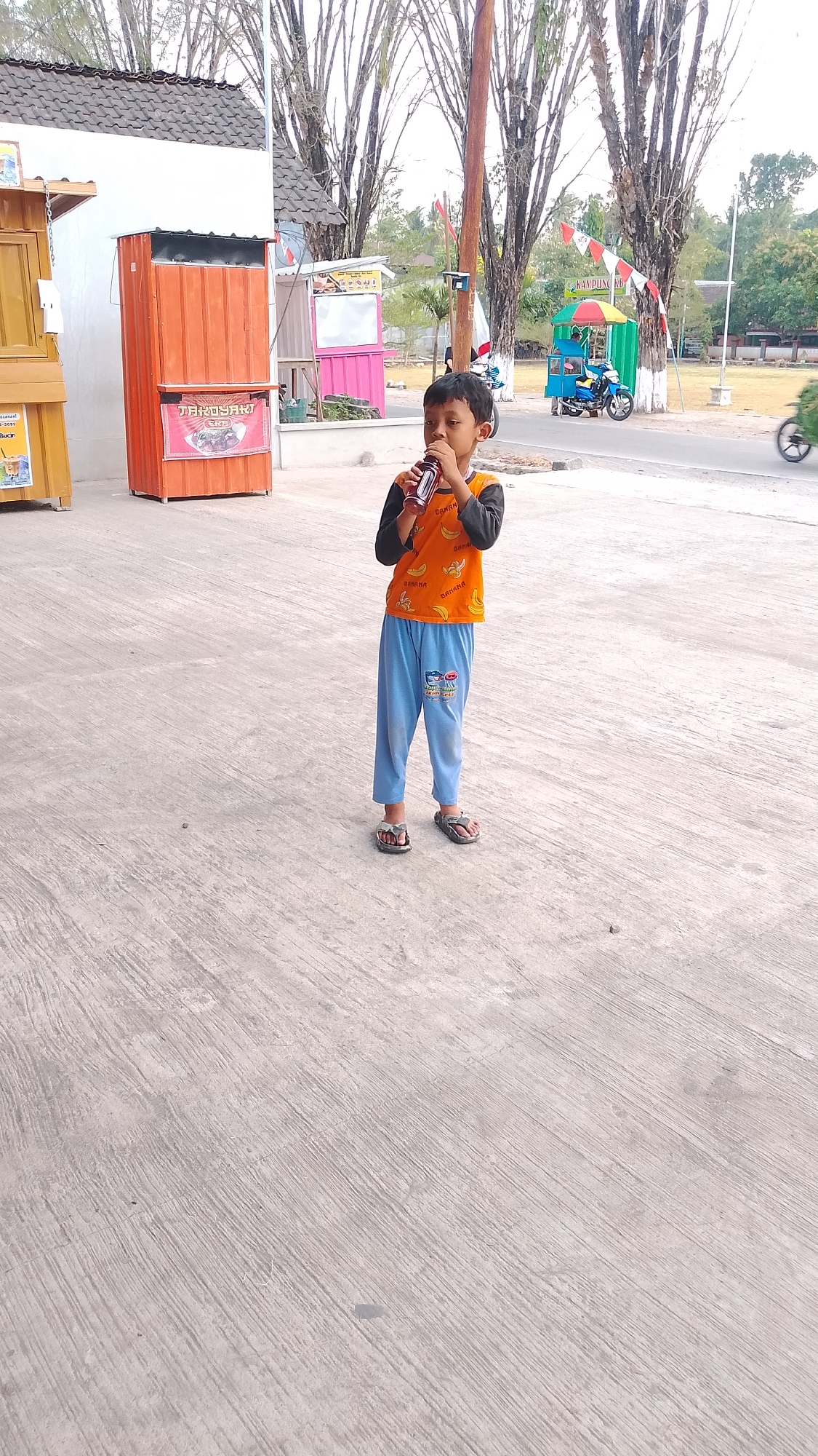 A young boy stands on a concrete surface, holding a bottle to his mouth. He wears an orange t-shirt with a banana print and blue pants. Behind him, to the left, is a food stall with an orange and brown facade. Further back, a pink structure can be seen, possibly another stall, with white signage. To the right, a street with a few parked motorcycles is visible, along with a red and white flag on a pole. The environment suggests an outdoor area, possibly a market or a roadside area in Karanglangu, Indonesia, with trees in the background. The overall scene is under daylight conditions.