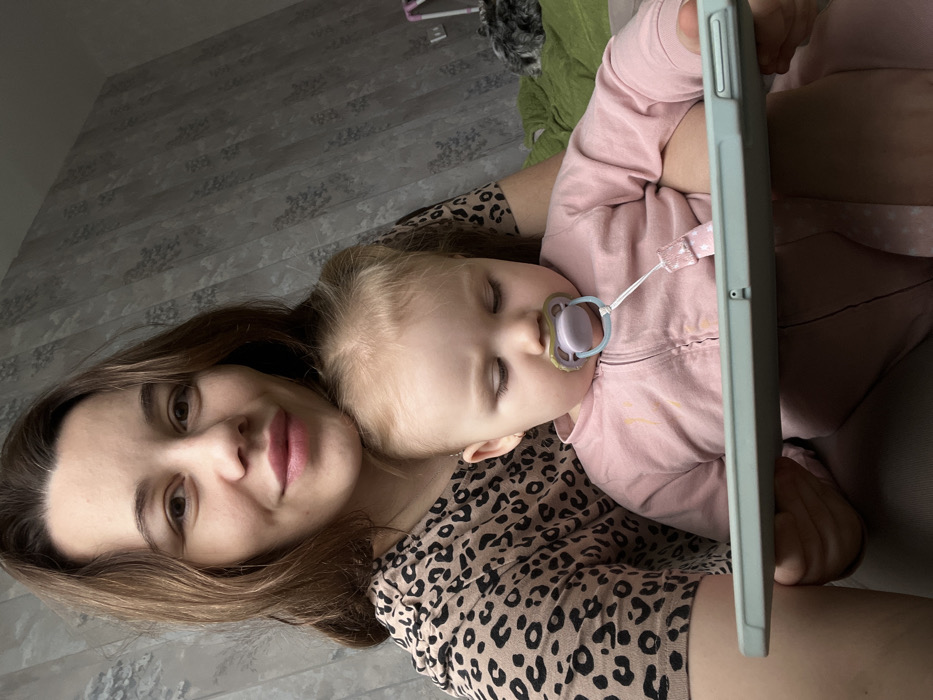 The image displays an adult female holding an infant indoors. The adult female, with brown hair, is positioned in the upper left, facing forward and smiling towards the camera. She is wearing a light brown and black leopard-print short-sleeved top. The infant, with light blonde hair, is seated in front of the adult, partially obscuring the adult's lower torso. The infant is wearing a light pink, long-sleeved, zippered romper with a few light-colored smudges on the front. A light purple pacifier with a clear ring and a pink strap featuring white stars is in the infant's mouth. The infant's eyes are directed downwards, focused on a light blue tablet enclosed in a protective case, which they are holding horizontally with both hands. The background consists of a wall covered in greyish-blue wallpaper patterned with subtle lighter grey and white floral or abstract designs. In the upper right corner of the background, a portion of green fabric, possibly a blanket or cushion, and a dark, furry object are visible. The specific location in the city of Leninakan, Russia, cannot be determined from the visual information provided within the image; Leninakan is the former Soviet-era name for Gyumri, Armenia.