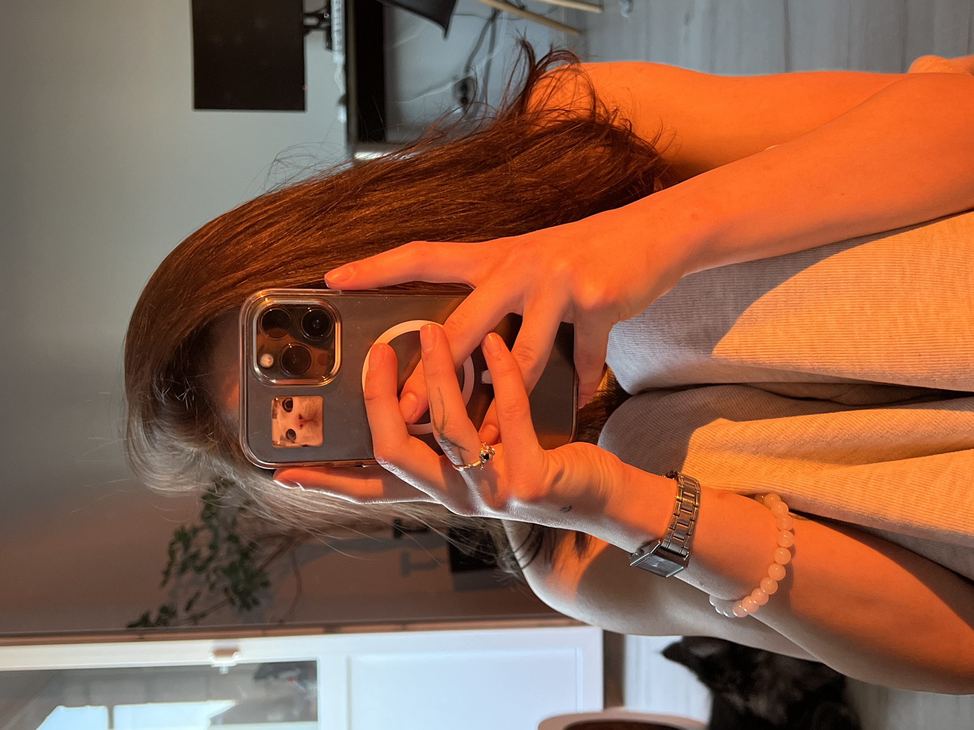 A person is taking a selfie with their phone. The phone has a small sticker of a cat's face on its case. The person's face is partially obscured by the phone. They are wearing a watch on their left wrist and a bracelet. In the background, there is a plant, a computer monitor, and what appears to be a pet lying on the floor. The lighting in the image is warm and orange, suggesting it was taken during sunset or sunrise. The image does not contain any identifiable landmarks or elements that would place it specifically in Ternopil, Ukraine.