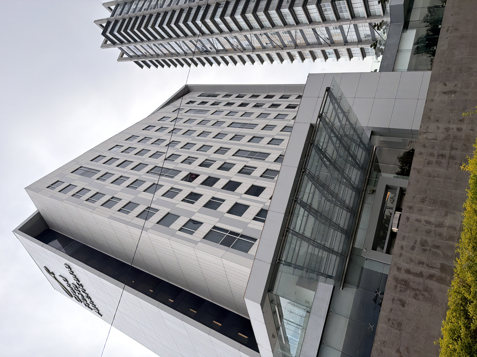 A low-angle shot captures two contemporary buildings in Puebla, Mexico. The primary building is a tall, rectangular structure with a gray-and-white facade punctuated by numerous windows arranged in a grid pattern. Its lower section features a dark horizontal stripe and a metallic logo. Adjacent to it is a glass-encased structure, revealing interior details and structural elements. At the top of the frame, a section of another modern building is visible, characterized by a series of layered, angular projections. To the right, a concrete wall and a green hedge partially obscure the view. The sky is overcast, providing soft, diffused light.