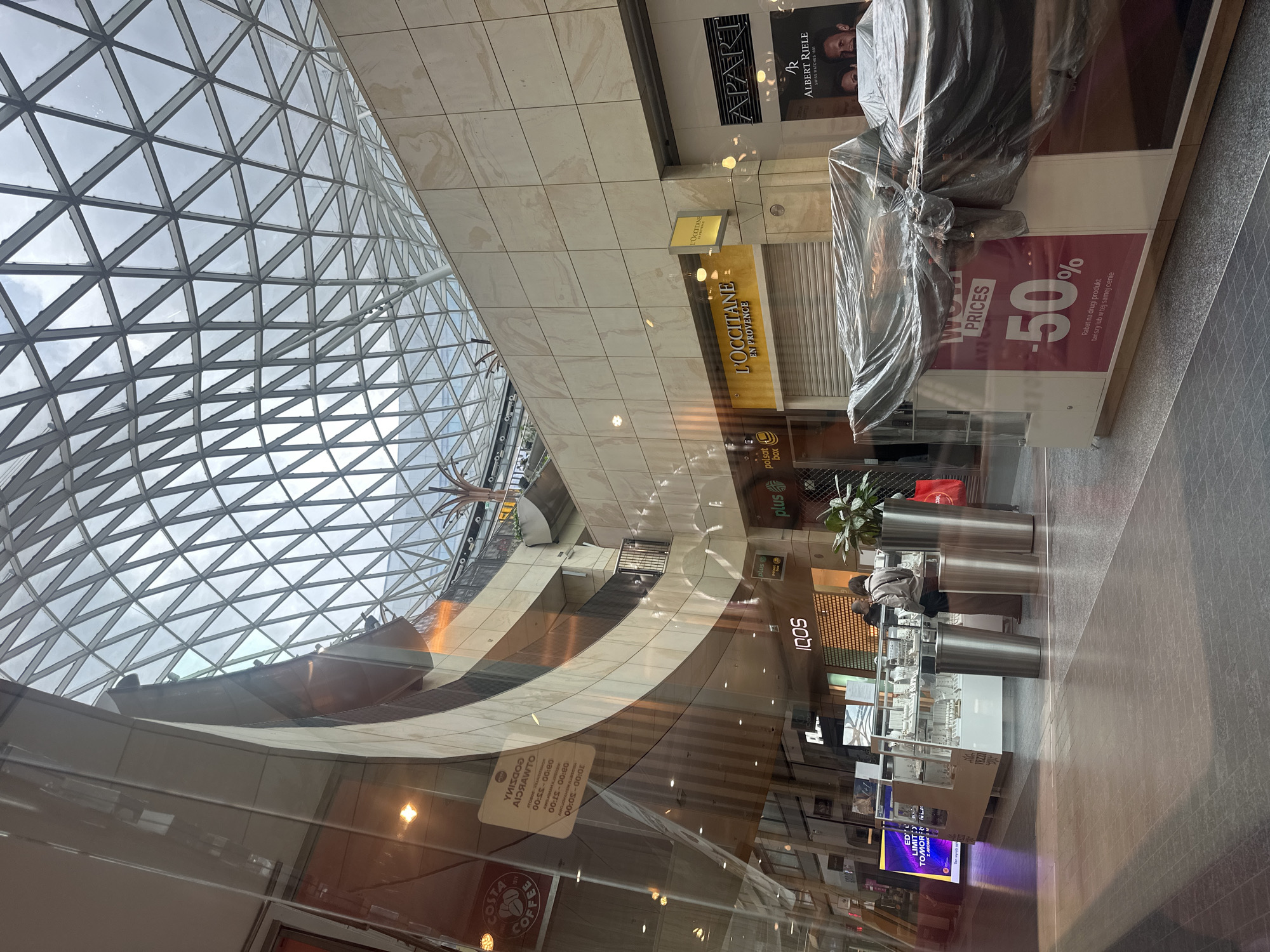 The image shows an indoor scene, likely a shopping mall, with a prominent glass and metal geodesic dome ceiling. The architecture features curved walls and multiple levels, with shops visible on the ground floor. One shop is labeled "L'OCCITANE EN PROVENCE," another is "ALBERT RIELE SWISS WATCHES 1891," and a "Costa Coffee" sign is also visible. There are also shops with names like "plus" and "IQOS." A jewelry display case is set up with a few customers browsing. Part of a display stand is covered with a tarp, displaying "WOW PRICES -50%". The floor is tiled in a dark gray pattern. The overall lighting suggests it is daytime, with natural light filtering through the dome. Based on the presence of the Polish language on signage, such as "Rabat na drugi produkt tańszy lub w tej samej cenie," the location is in Warsaw, Poland.