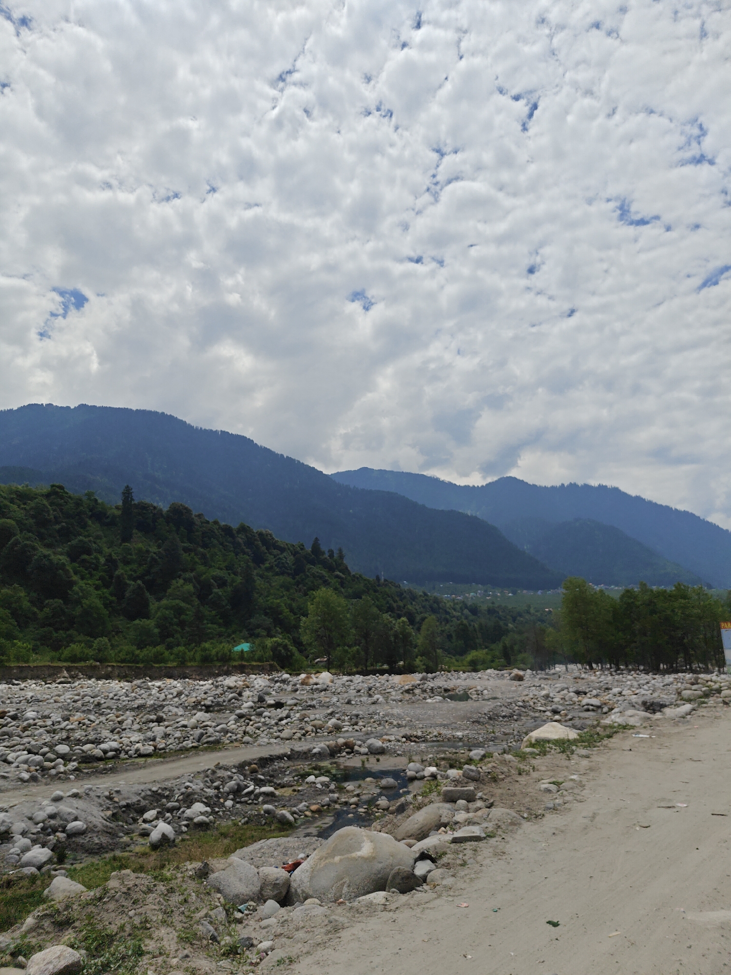 This outdoor landscape photograph captures a wide, serene view of a mountainous region in Kalath, India. The dominant feature is a vast expanse of light grey and white clouds scattered across a mostly bright sky, with glimpses of blue visible between them, suggesting an overcast or partly cloudy day. The light is diffused, indicating it's daytime, likely late morning or afternoon.
In the middle ground, majestic, dark green mountains rise prominently, covered in dense, temperate forest. Their peaks are shrouded in a light haze, contributing to the sense of distance and scale. Below the mountains and extending into the foreground is a wide, rocky riverbed, currently dry or with very low water levels, revealing countless smooth, light-colored boulders and pebbles. A narrow, lighter dirt track or path traverses the rocky bed from the left.
On the immediate right of the foreground, a sandy dirt road or path runs alongside the riverbed, dotted with scattered small rocks and some debris. Patches of green grass and small shrubs grow intermittently along the edges of the dirt path and within the rocky riverbed. On the left side of the riverbed, beyond some sparse vegetation, a small, vibrant green roof of a structure is barely visible. In the far distance, nestled between the mountain ranges, a small clustered settlement or village can be seen. To the far right, partially obscured by trees, a sign with the letters "PARA" visible on a yellow background suggests a local business, possibly related to tourism like paragliding. No people are visible in the scene. The overall impression is one of natural grandeur and tranquility.