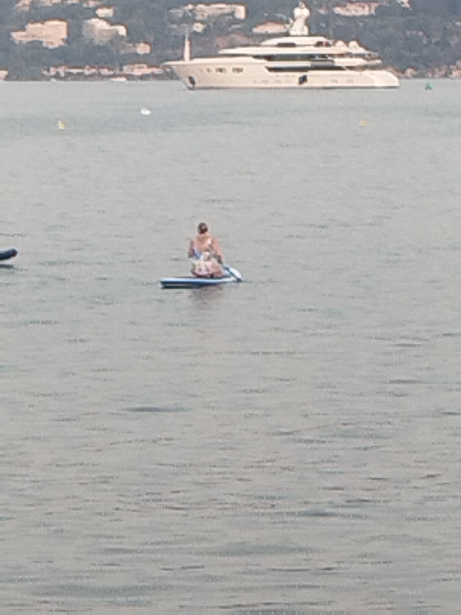 The image captures a calm body of water, likely the Mediterranean Sea, near Antibes, France. In the background, a large white yacht is moored, with buildings and lush green hillsides forming the coastline. In the foreground, a person is seated on a blue and white paddleboard, holding a paddle. The water is a muted gray-blue, with subtle ripples. The overall scene suggests a leisurely day at sea.
