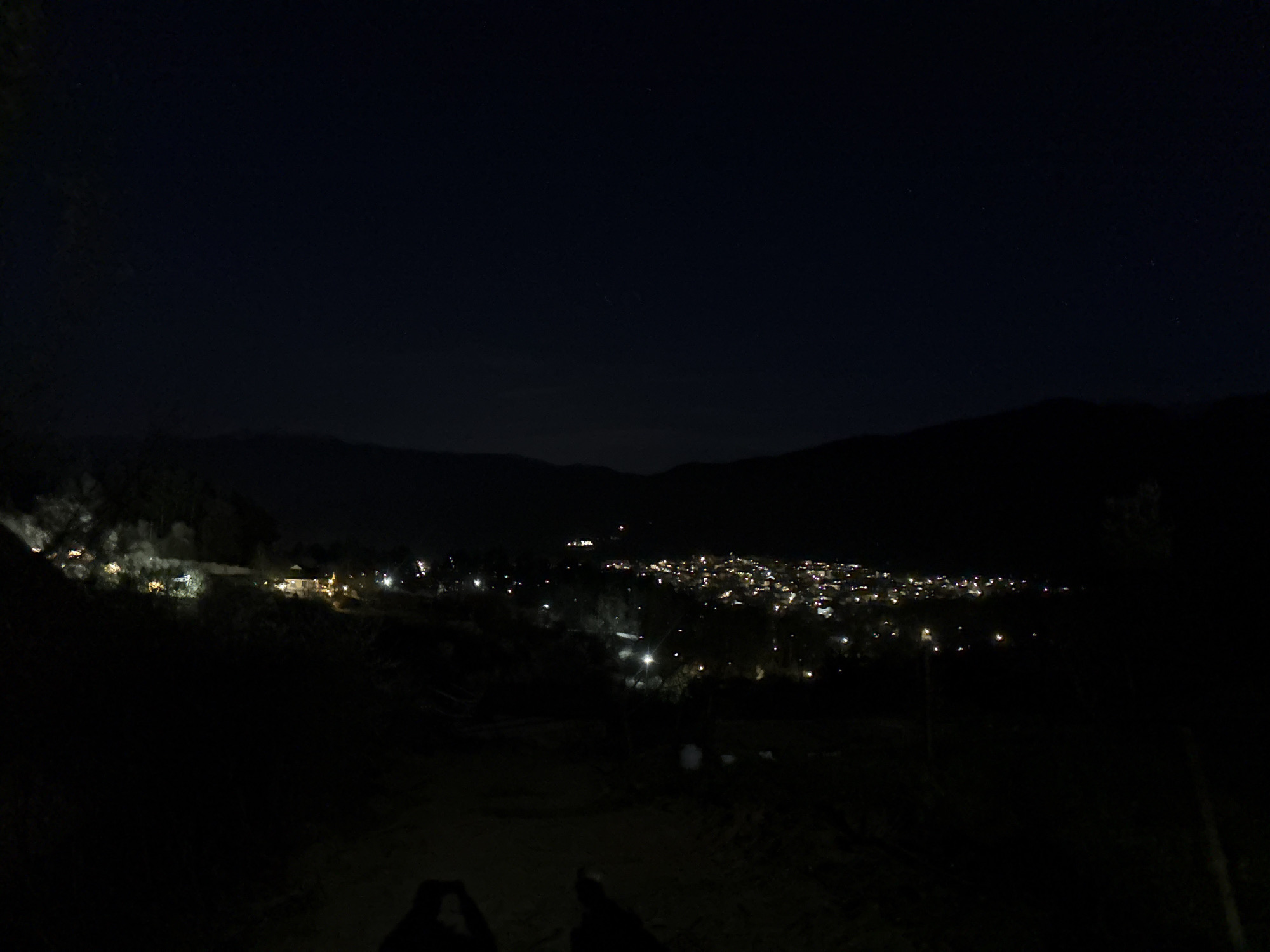 The image shows a nighttime view of Berkovitsa, Bulgaria, from an elevated perspective. The city lights are visible in the valley, forming a cluster of illumination against the dark landscape. Silhouettes of mountains are seen in the background, with a dark night sky above. The foreground is obscured in darkness, with indistinct shapes possibly indicating ground or objects. The scene is quiet and still, with the city lights being the primary visual element.