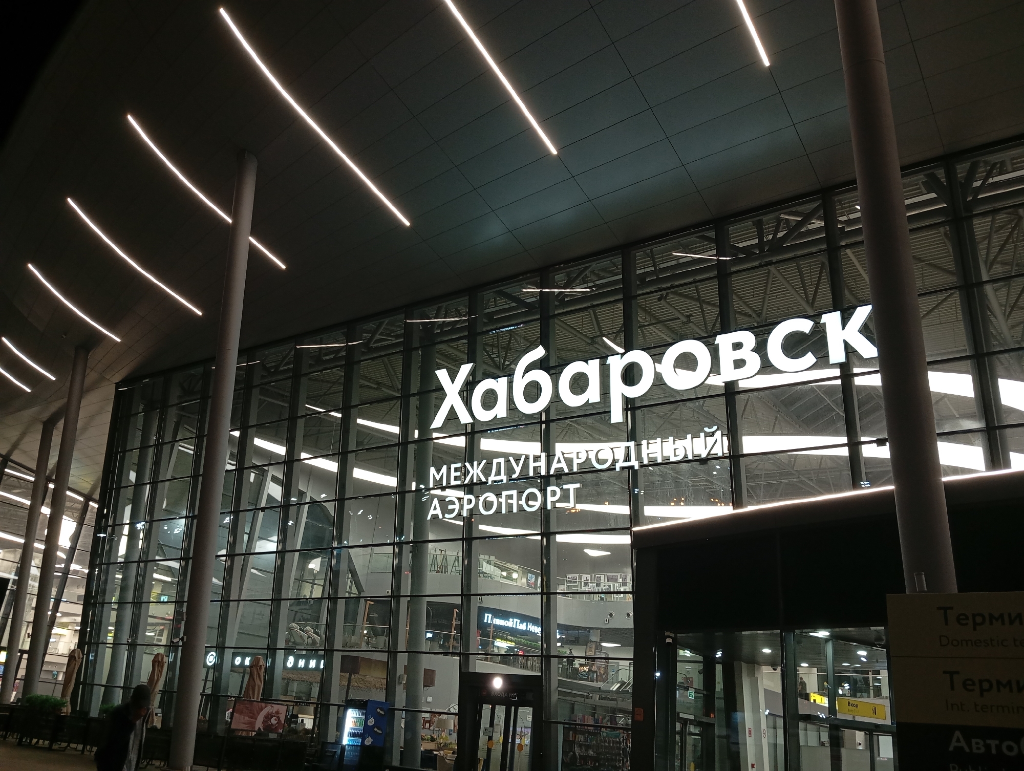 The image depicts the exterior of the Khabarovsk Novy Airport in Khabarovsk, Russia. The airport terminal features a large, modern facade made of glass and metal. Prominently displayed in large white lettering on the glass are the words "ХАБАРОВСК" (Khabarovsk) and below that, "МЕЖДУНАРОДНЫЙ АЭРОПОРТ" (International Airport). The building is illuminated by a series of long, linear lights installed on the dark, angled ceiling above the entrance. Several tall, cylindrical columns support the roof structure. Outside the terminal, there are some outdoor umbrellas and a vending machine visible. The scene appears to be captured at night or during low light conditions, with artificial lighting being the primary source of illumination. The reflection of the interior lights and architectural elements is visible on the glass facade.