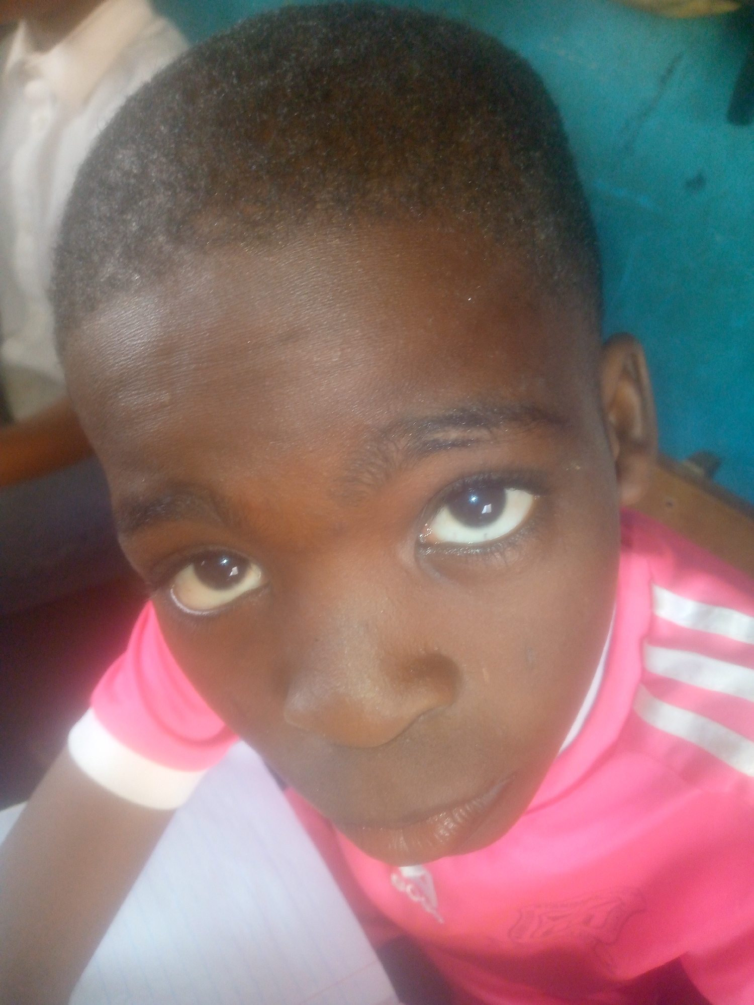A close-up shot focuses on a dark-skinned child wearing a pink shirt with white stripes. The child appears to be indoors, potentially sitting at a desk with a piece of paper visible. The location is unknown.