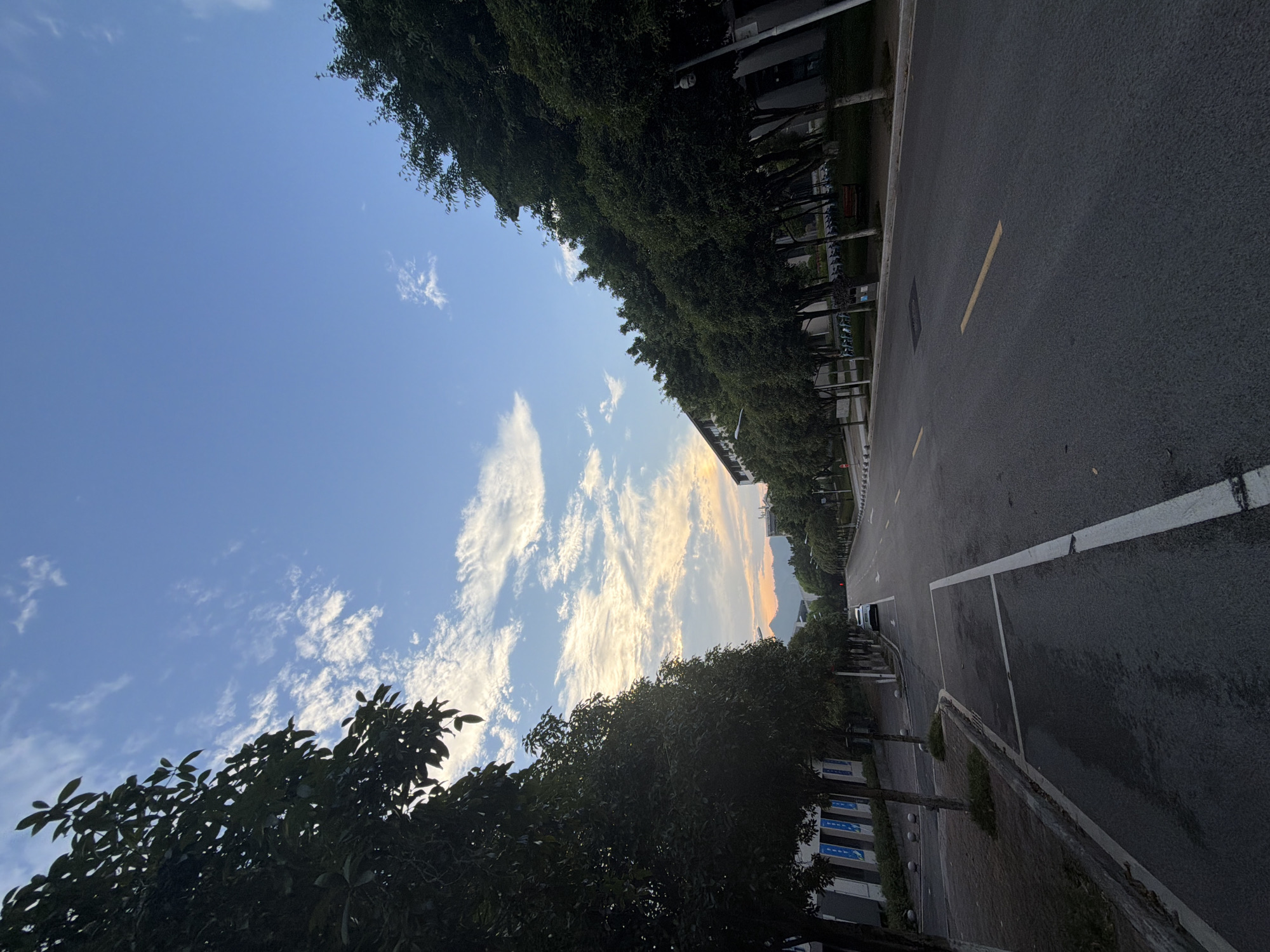 The image depicts an exterior view of a street lined with trees and buildings, likely in the city of Jiangjin, China. The scene is captured during what appears to be dawn or dusk, with the sky showing a gradient of blue and wispy clouds, illuminated by the soft glow of sunrise or sunset near the horizon, casting a warm light on distant mountains. On the left side of the street, a sidewalk is bordered by trees and a low building with blue signage. The street itself is asphalt with yellow and white lane markings. A white car is parked on the right side of the street, further down, adjacent to a row of trees and a building complex. The environment appears calm and devoid of prominent human activity at this moment.