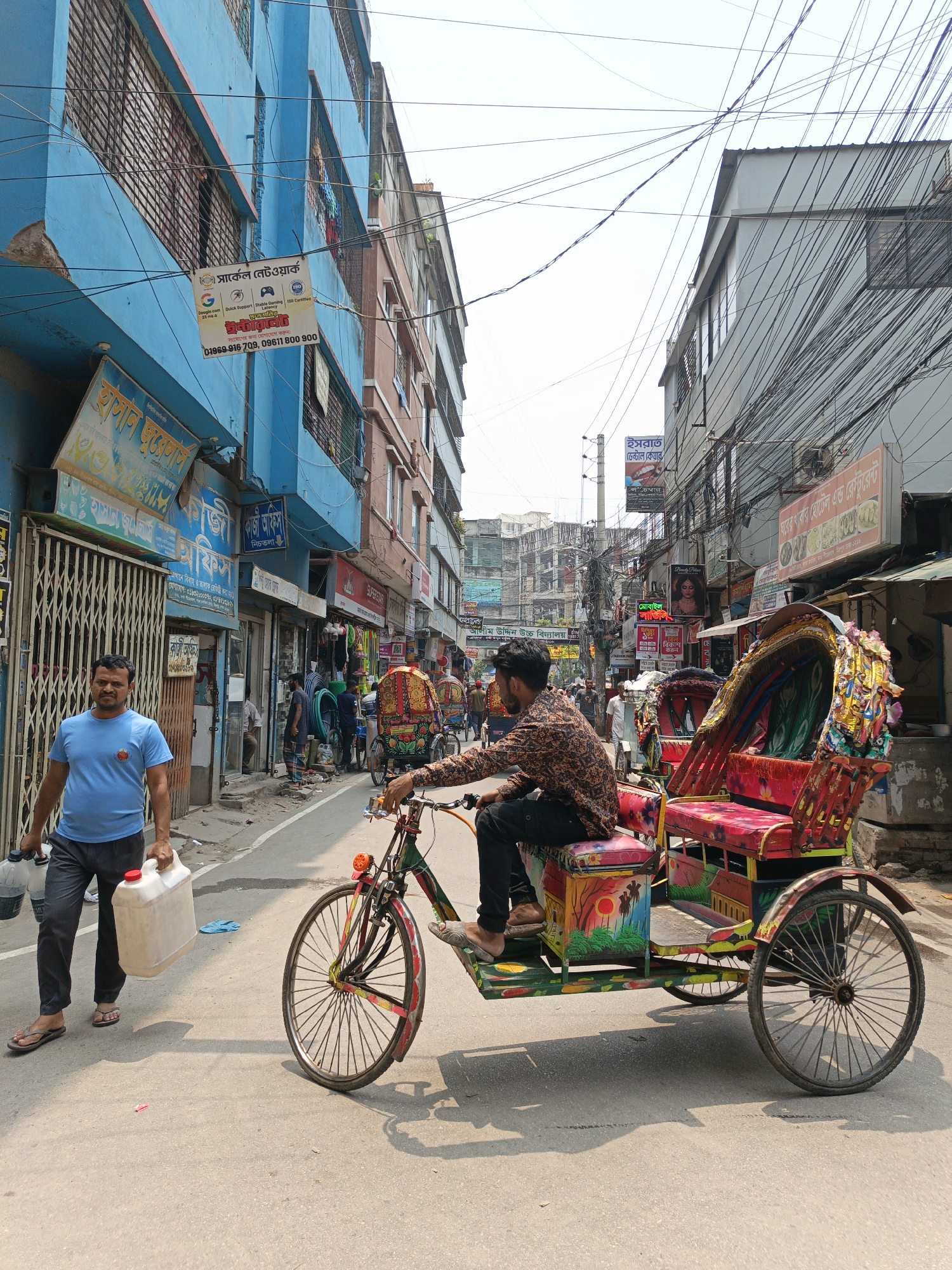 The image shows a street scene in what appears to be a densely populated urban area, possibly in South Asia, given the rickshaws and signage in a language other than English. The street is paved and relatively narrow, flanked by buildings of varying heights and colors. Overhead, a dense network of electrical wires creates a chaotic pattern against the sky.
A man in a blue t-shirt and dark pants walks along the left side of the street, carrying white plastic containers. To his right, a man is riding a brightly colored rickshaw. The rickshaw is intricately decorated with vibrant colors and designs. Several other people are visible in the background, walking along the street.
The buildings are a mix of blue, brown, and grey, with various shops and businesses at street level. Signboards with writing in a non-English language are visible above the storefronts. The overall impression is of a busy, vibrant, and somewhat chaotic urban environment.