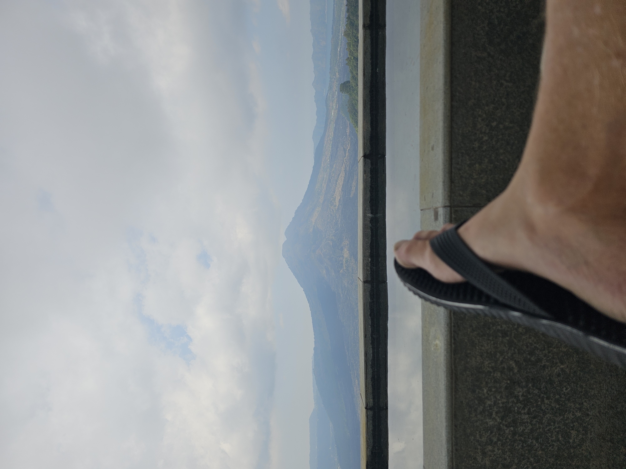 The image, taken in Kintamani, Indonesia, presents a first-person perspective of a person relaxing near an infinity pool. The foreground is dominated by the person's right foot, clad in a black flip-flop, resting on the edge of the pool. The pool's surface reflects the cloudy sky above. Beyond the pool, a tiered landscape unfolds, characterized by rolling hills and mountains shrouded in a hazy atmosphere. The most prominent feature is a large, conical mountain rising majestically in the mid-ground, its slopes displaying varying shades of green and brown, indicative of vegetation and possibly volcanic rock. The sky is overcast with soft, diffused light. The overall impression is one of tranquility and natural beauty, enhanced by the presence of the person's relaxed posture.