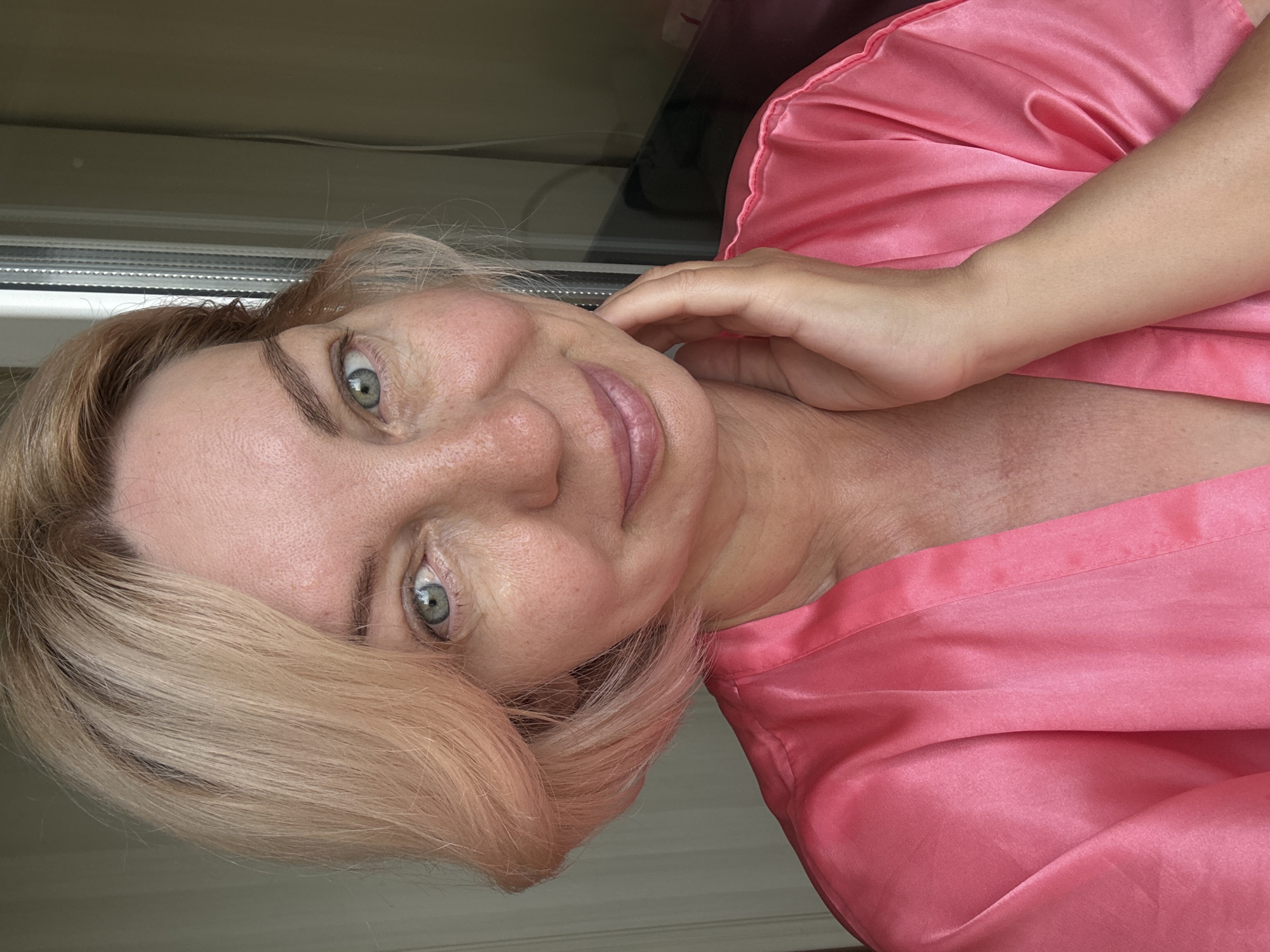 The media file is a close-up, vertically oriented portrait of a woman with fair skin and light blonde hair. She is wearing a silky pink robe, which is draped over her shoulder and chest. Her left hand is gently touching her cheek, and her head is tilted slightly. She has light blue-green eyes and is wearing subtle makeup, including lipstick in a muted pink shade. Her expression appears relaxed and serene. The background is out of focus, but hints of what appear to be blinds or a window frame are visible, suggesting an indoor setting near a window. The lighting suggests it might be daytime, possibly late afternoon given the soft quality of the light. There is no visible text in the image. The context provided indicates the location is Sochi, Russia.