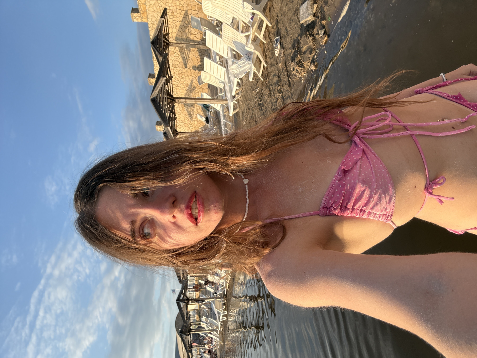 This is a portrait of a young woman posing for a selfie in the Dead Sea, located in Sol-Iletsk, Russia. The woman has long, brown hair and is wearing a pink polka dot bikini. She has makeup on, with visible lipstick. She is in the water, which appears to be dark and murky. In the background, there is a beach with white lounge chairs and umbrellas. There are also stone buildings and wooden structures that appear to be part of a resort or hotel. The sky is blue with some scattered clouds, suggesting it is daytime, likely late afternoon or early evening due to the warm lighting. The overall mood appears relaxed and happy. There is no visible text in the image.