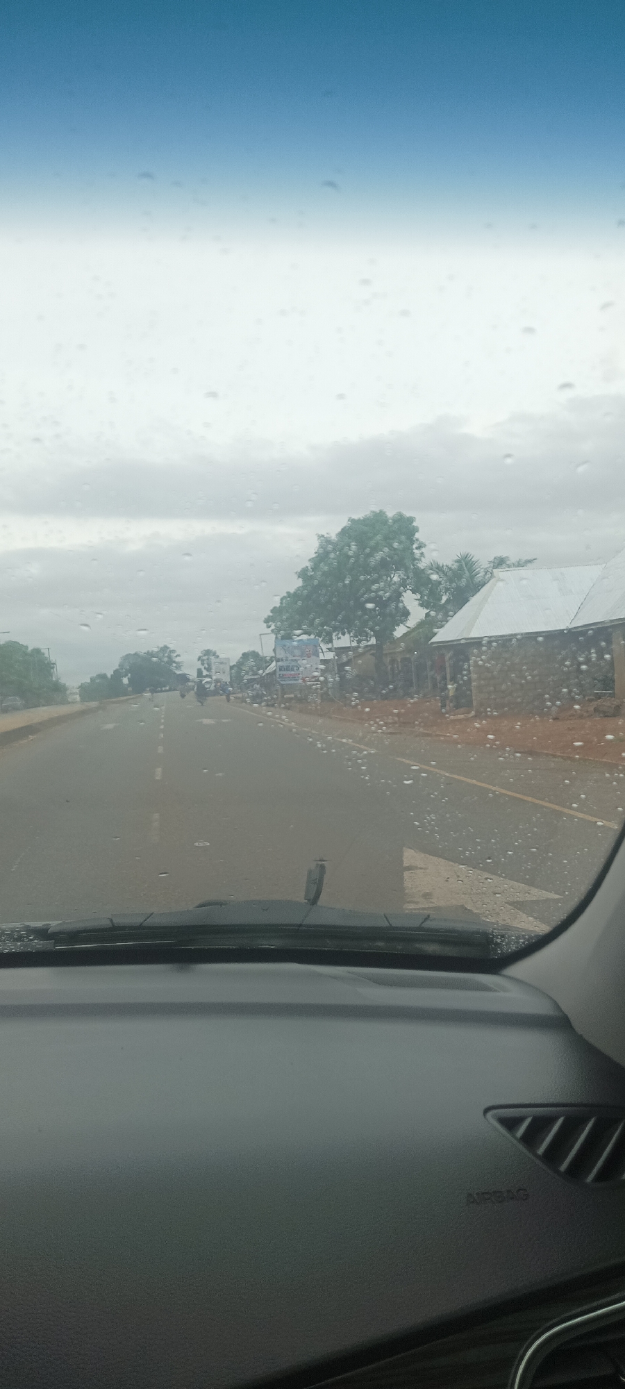 The image presents a view from inside a vehicle traveling on a wet, paved road, likely in the vicinity of Gwadanye, Nigeria, based on the metadata. The windshield is covered in raindrops. The road has white dashed lane markings and an arrow marking. In the distance, there are several motorcycles and buildings along the road, including some with corrugated metal roofs. There is a billboard visible near a tree. The sky is overcast. The interior of the vehicle is visible in the foreground, including the dashboard and the "AIRBAG" label.
