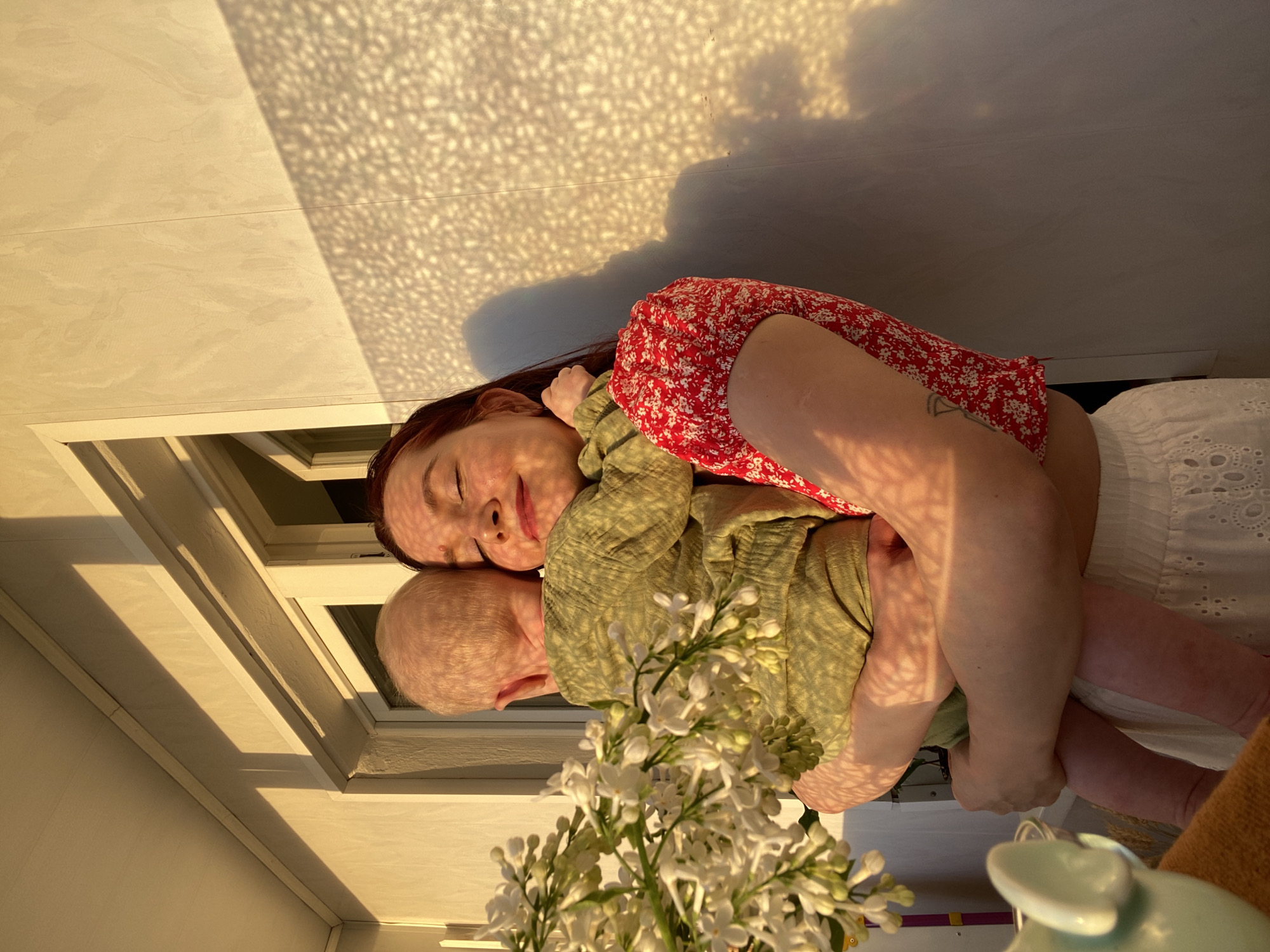 A woman with red hair, wearing a red floral top and a white skirt, is holding a baby in a light green shirt. The baby has a bald head. The woman is lying on a surface, possibly a bed or floor, next to a white window. A bouquet of white flowers is in the foreground. Sunlight is casting a patterned shadow on the wall behind the woman and baby. The scene appears to be indoors, likely in a home. There is no indication that the image was taken in Chaikovsky, Russia.