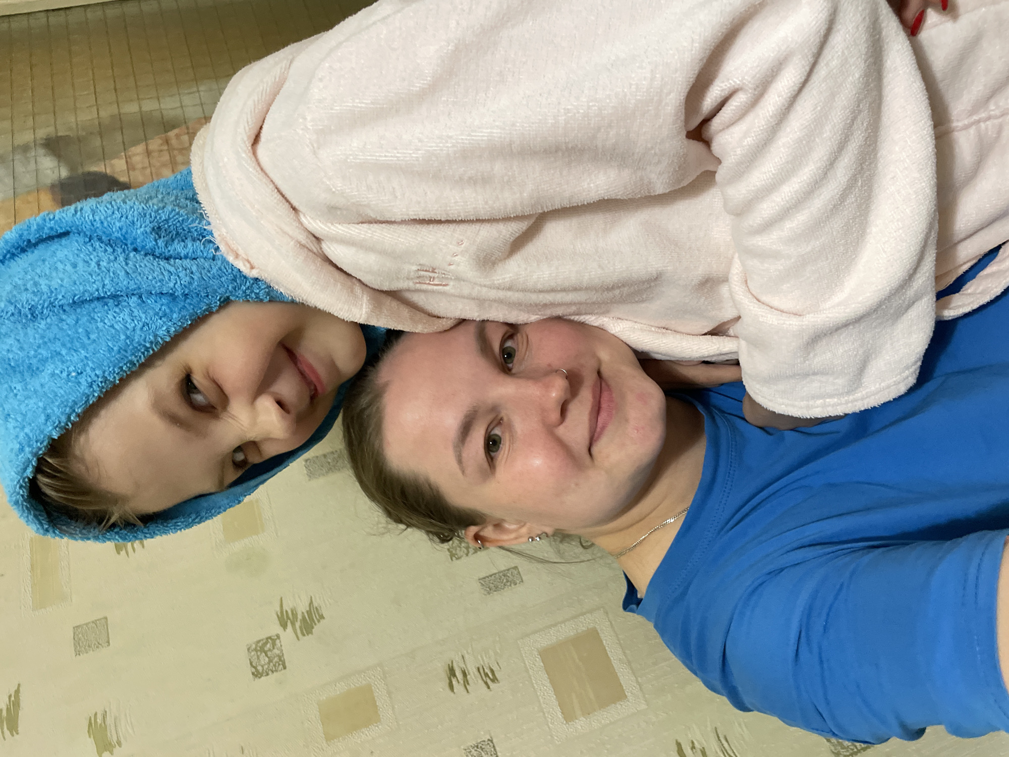 A close-up, medium-shot photograph shows two individuals, possibly a mother and child, lying on a light-colored surface with a patterned design. The person in the lower right of the frame is wearing a blue T-shirt and a silver necklace and has multiple earrings. The other individual, possibly a child, is positioned above them; they are wearing a light pink bathrobe and a blue towel on their head. Both individuals are looking directly at the camera with smiles on their faces. The background is out of focus. Based on available visual cues, the location cannot be determined as Izhevsk, Russia.