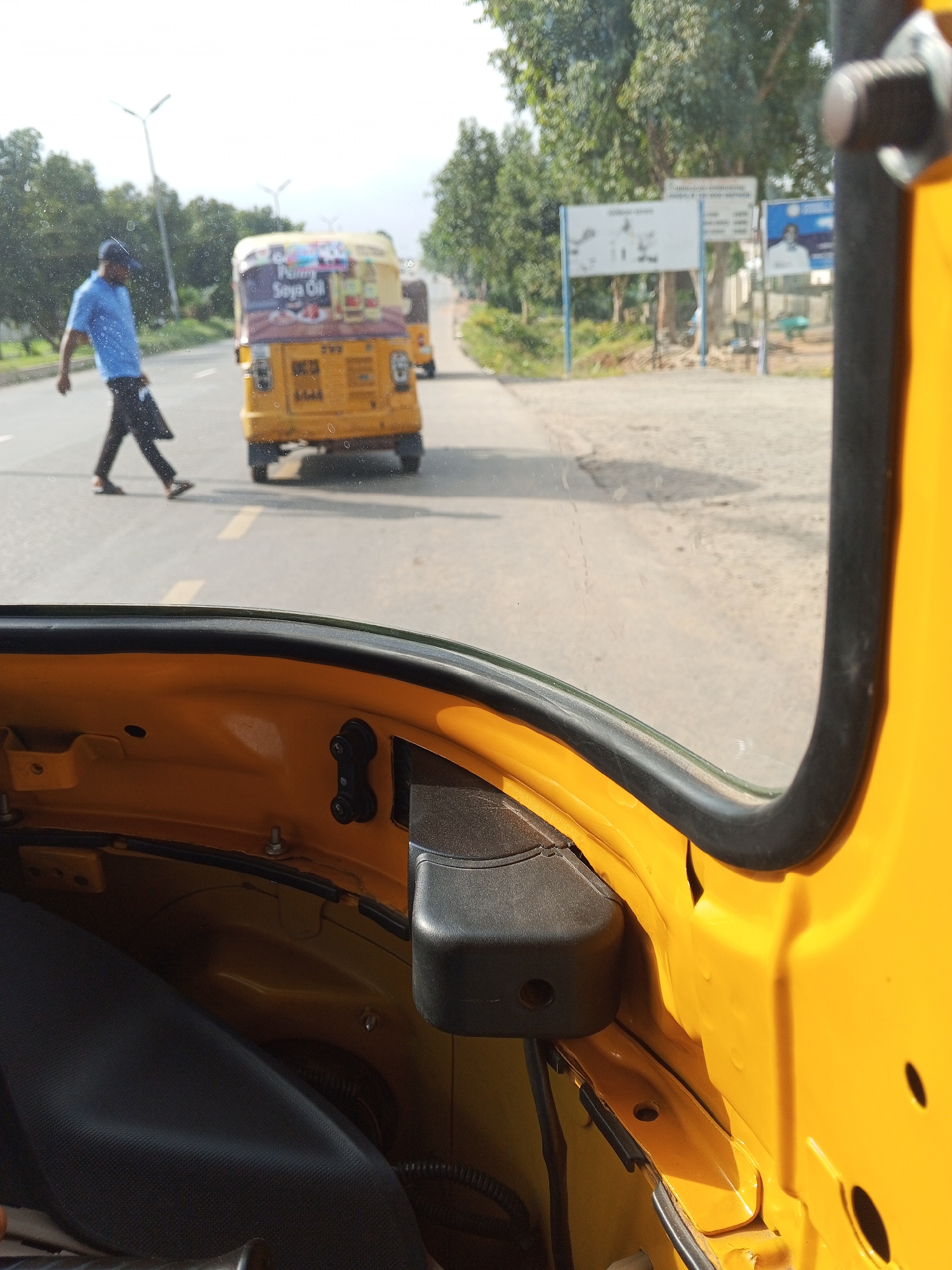 The image captures a street scene in Abuja, Nigeria, from the perspective of a passenger inside a yellow auto-rickshaw. A man in a blue shirt and cap is walking across the road, facing left. Behind him, a yellow auto-rickshaw with advertisements on its side is driving away. Further down the road, another auto-rickshaw is visible. The environment appears to be a typical roadside with trees and streetlights in the background. The foreground shows the interior of the auto-rickshaw, with its yellow paint and black seating. The image is taken during daylight.