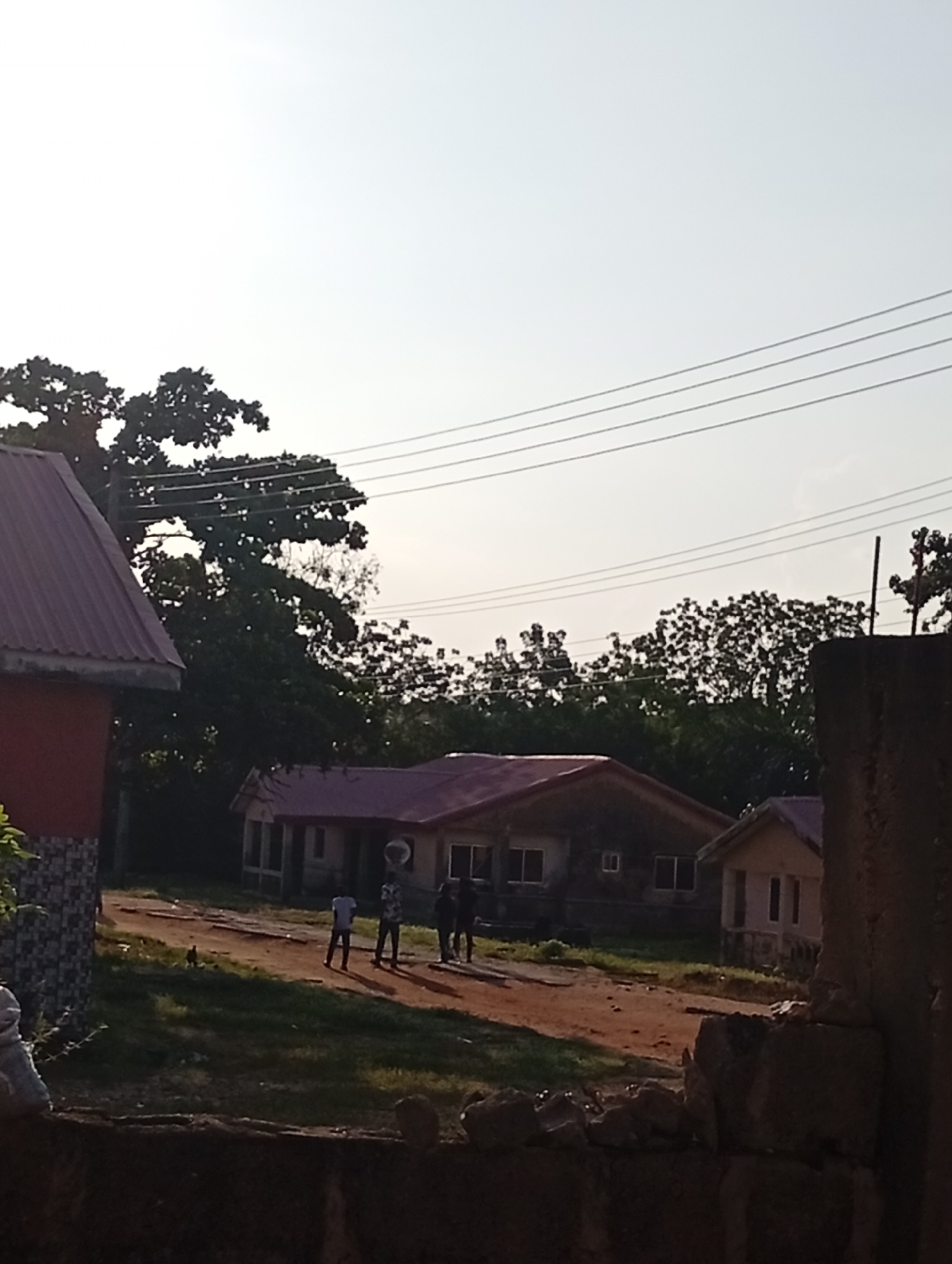 In Adankolo, Nigeria, an outdoor scene unfolds with a clear sky and scattered trees. In the foreground, a partially visible brick structure with rough-hewn stones forms a boundary. Beyond this, a grassy area leads to a dirt path where four individuals are visible. Three of them are standing together, appearing to engage in conversation or observe something in the distance. One person is slightly set apart, facing away from the others and carrying an object on their head. Behind the figures, a low-slung building with a red roof stands, its windows visible. Further in the background, another smaller building with a similar red roof is present. The environment is characterized by a dusty ground, sparse vegetation, and overhead power lines supported by poles. The overall impression is of a residential or communal area during daylight hours.