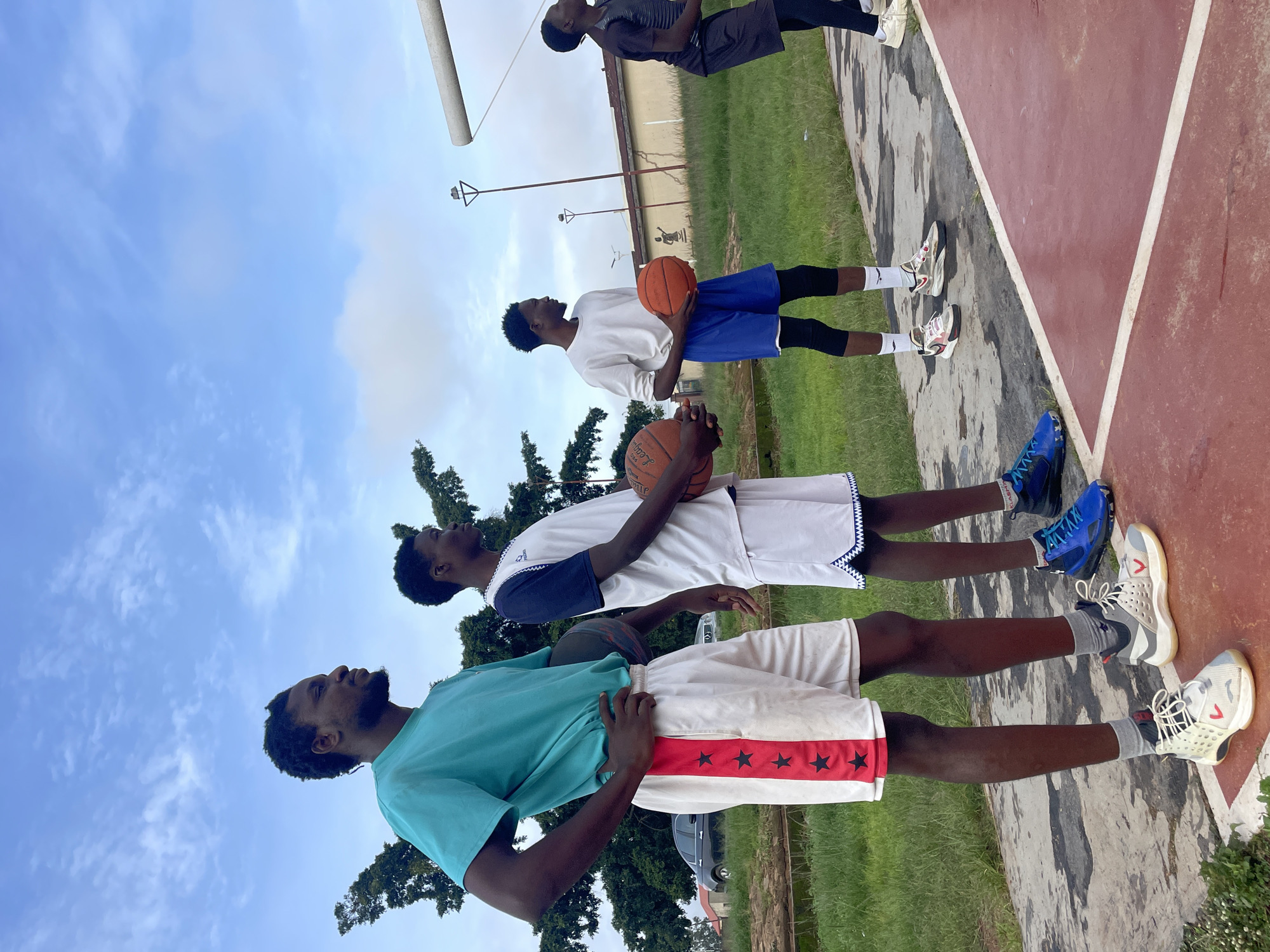 Four young Black men stand on a basketball court, holding basketballs. The man on the far left wears a teal t-shirt and white shorts with red stripes and black stars. The man next to him wears a white and blue jersey with white shorts. The third man from the left wears a long-sleeved white shirt and blue shorts. The man on the far right wears a dark gray sleeveless shirt and black shorts. They are outdoors with a cloudy blue sky above. In the background, there are trees, buildings, a street light, and a basketball hoop. The court surface is red and gray. There is grass and a fence in the background.