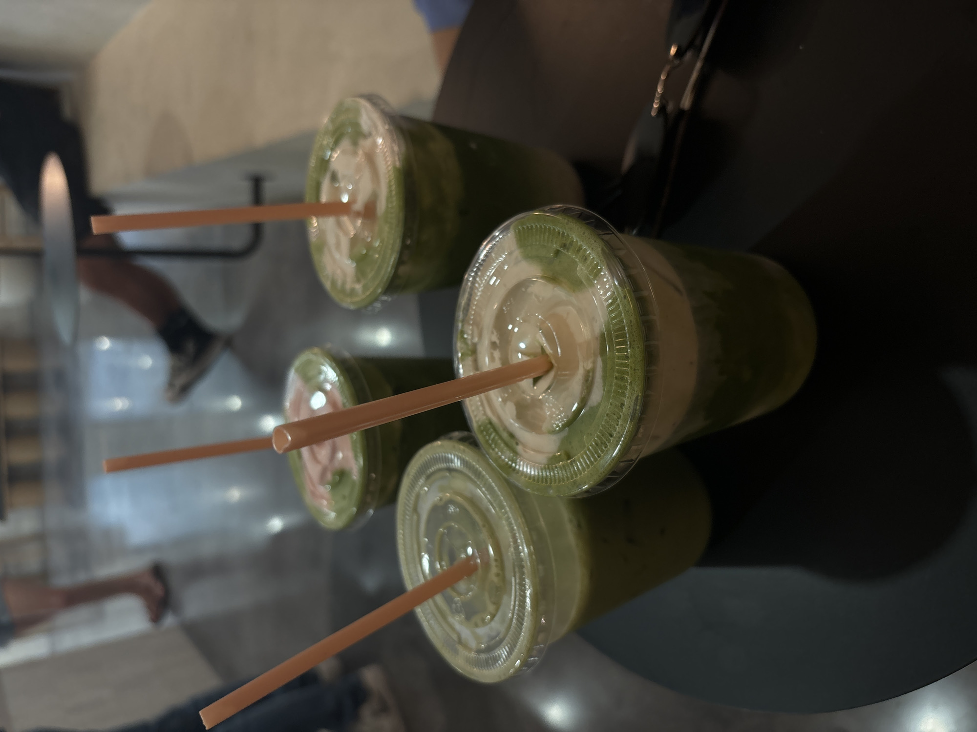 Four iced beverages are presented in clear plastic cups with domed lids and orange straws. Three of the cups are positioned in the foreground, angled slightly towards the viewer, and are filled with green liquids and some creamy white topping. The fourth cup, further back and to the right, also contains a green beverage. The beverages appear to be a matcha latte or smoothie. The cups sit on a dark, circular table. In the blurred background, two individuals are partially visible; one on the left is wearing shorts and sandals, and another on the right is seated, with their legs crossed. The environment suggests an indoor cafe or restaurant setting. The overall lighting is dim, with some highlights on the table and beverages. The location is identified as Hollywood, United States of America.