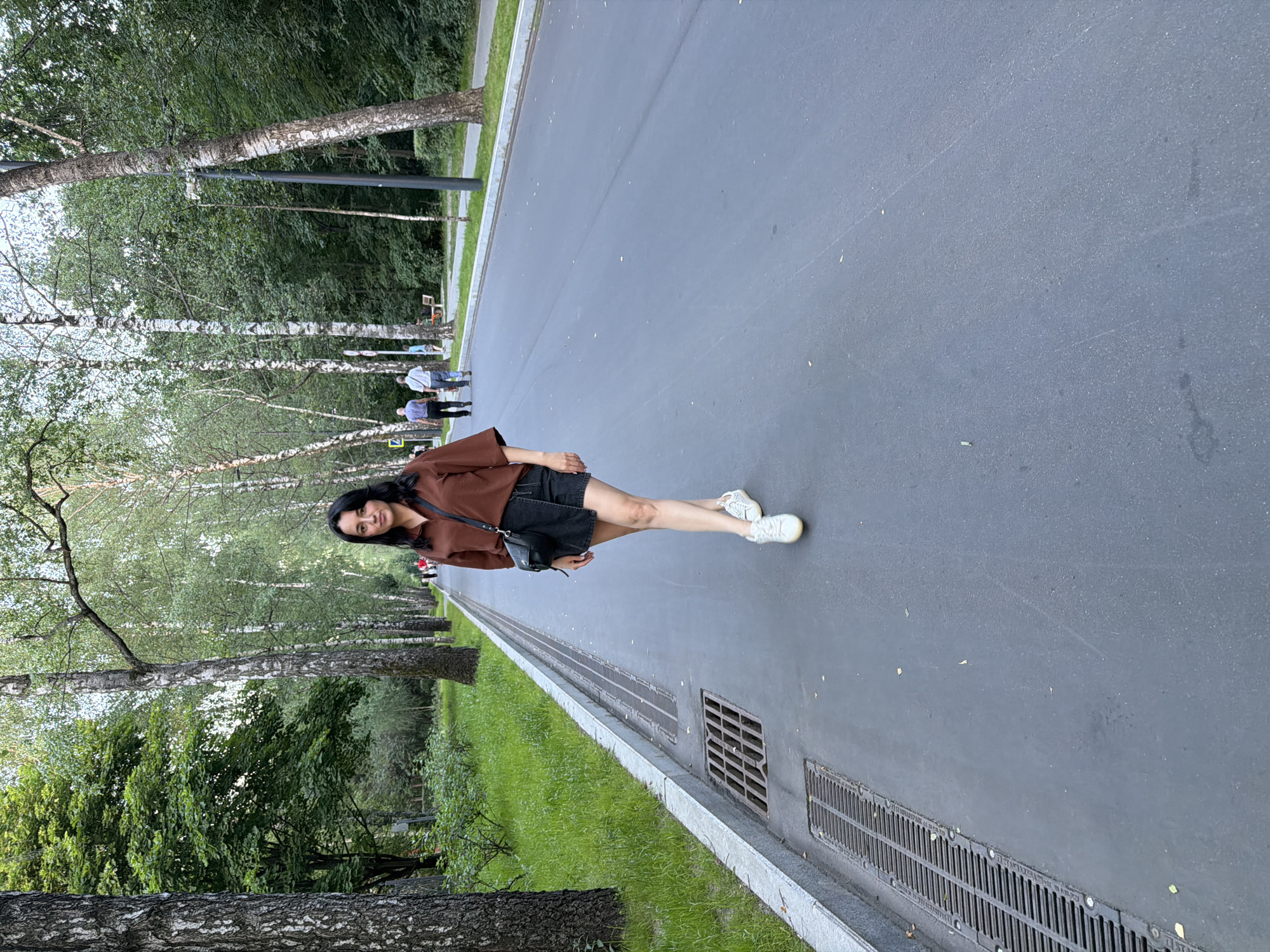 A woman with dark hair, wearing a brown collared shirt, a black asymmetric skirt, and white sneakers, walks towards the camera on an asphalt path. She carries a black cross-body bag. The path is lined with trees, predominantly birch trees, with green grass and foliage on the left. In the background, other people are walking along the path. This scene appears to be in a park in Moscow, Russia, possibly Sokolniki Park, given the characteristic tree-lined paths and the general urban park aesthetic. The lighting suggests it is late afternoon or early evening.