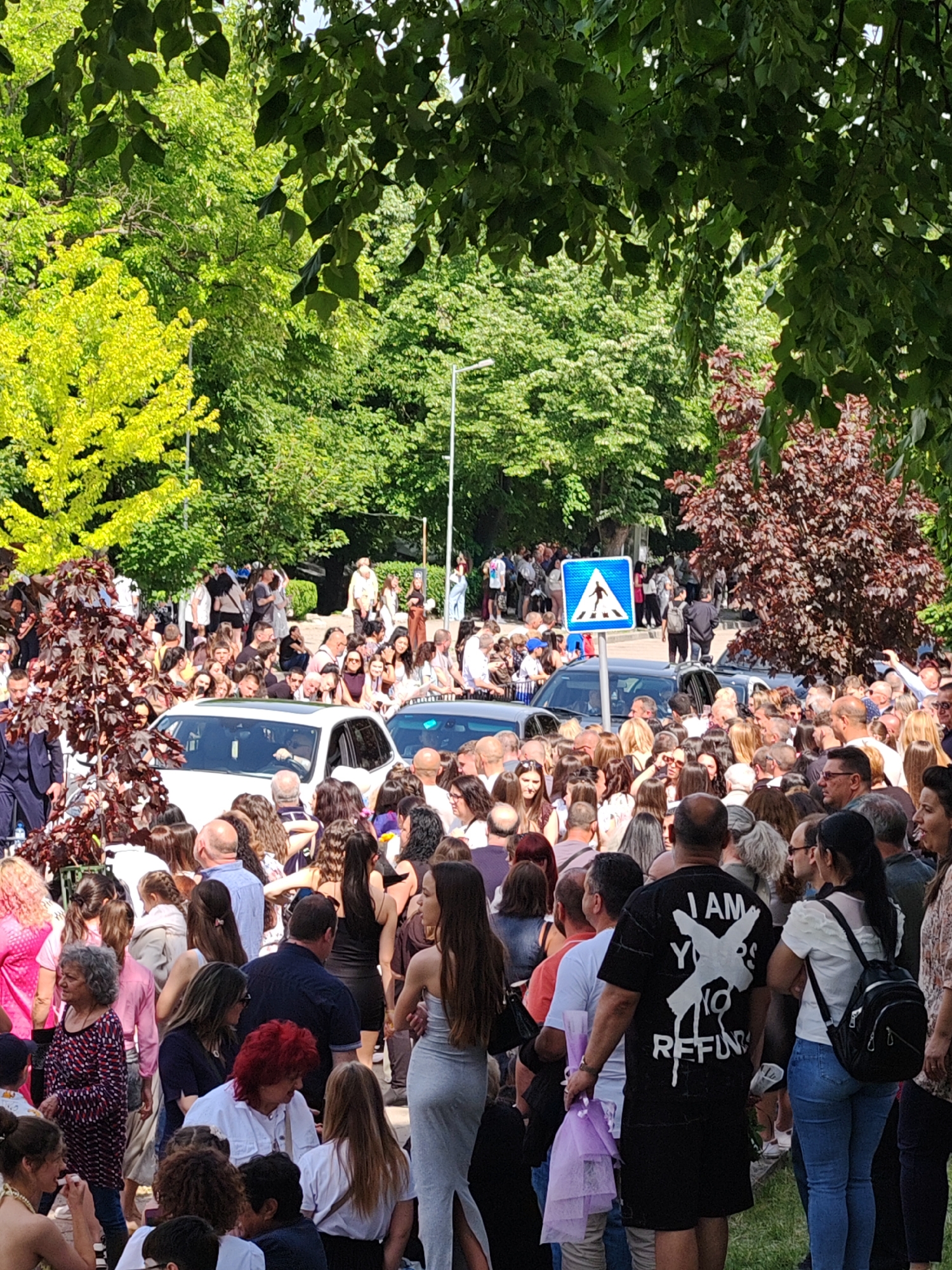 The image shows a dense crowd of people gathered outdoors, likely in a public area in Shumen, Bulgaria. The environment features trees and other greenery typical of a park or urban garden. The crowd consists of men and women, with varied hairstyles and clothing, suggesting a diverse demographic. A pedestrian crossing sign is visible in the background, adjacent to a parked vehicle, suggesting the crowd is near a road or crosswalk. The overall scene conveys a sense of an event, celebration, or public gathering. The image captures a moment of potential interaction among many people. The specific purpose of the gathering is not evident from the image alone.