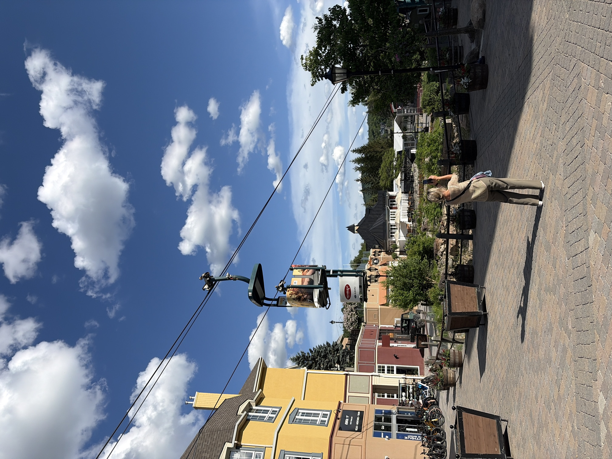 In Mont-Tremblant, Canada, a woman stands on a brick pathway, holding a smartphone up to take a photograph. She is dressed in olive green pants and a light brown, short-sleeved shirt, and she is facing away from the camera. Behind her, a ski lift gondola hangs suspended from a cable, displaying advertisements on its sides. The gondola is moving across the frame from left to right.
The scene is set in a village-like area with buildings constructed in a European architectural style, painted in vibrant colors like yellow and orange, with dark-colored roofs. The pathway is lined with decorative planters, some filled with flowers, and wooden benches. In the background, more buildings, trees, and rolling hills can be seen under a bright blue sky dotted with fluffy white clouds. A black SUV is parked near a building on the left side of the image, and several bicycles are leaning against a storefront. The lighting suggests it is a sunny day.