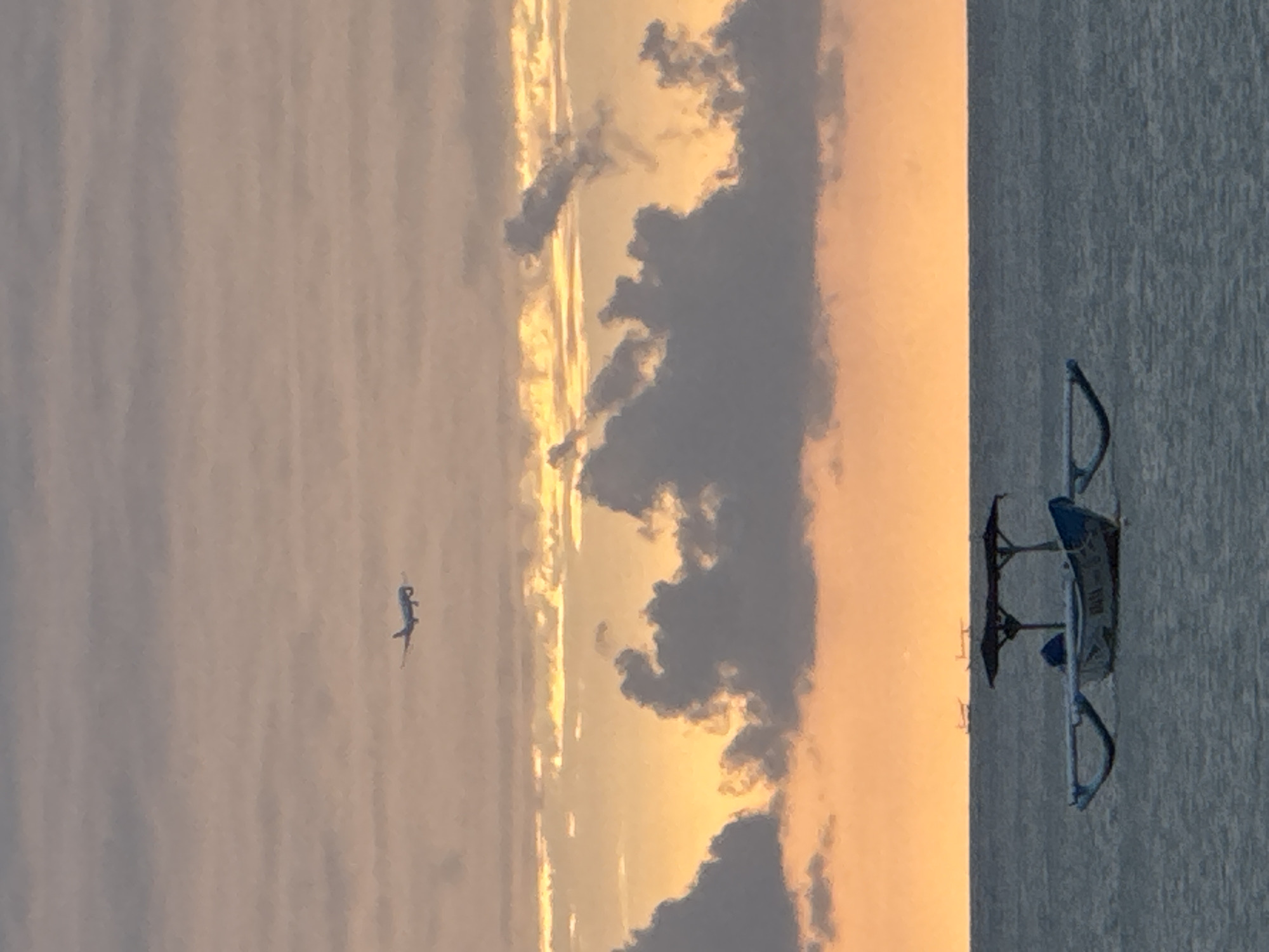 An airplane is flying in the sky over the ocean at sunset. The sky is filled with clouds that are lit up by the setting sun. In the foreground, there is a traditional Indonesian outrigger boat floating on the water. The boat is blue and white and has a canopy over the middle. The ocean is calm and reflects the colors of the sky. This scene likely takes place in Kuta Selatan, Indonesia, a popular tourist destination known for its beaches and sunsets.