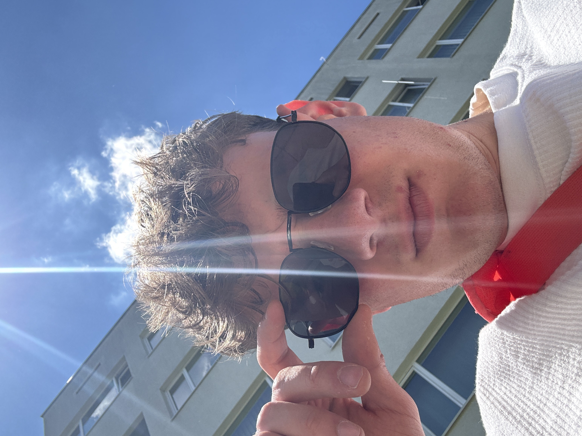 A low-angle, selfie-style photograph depicts a fair-skinned male wearing dark sunglasses, touching the lower rim of the eyewear with his left hand. He has short, curly brown hair. He is wearing a white, textured garment with a red strap or accent visible at the neck.
Behind the man, a modern building with multiple windows is visible. The sky is blue with scattered clouds and a bright sun, causing lens flare.
The location cannot be determined from the image. There is no information present in the photograph that suggests that the location is Sofia, Bulgaria.