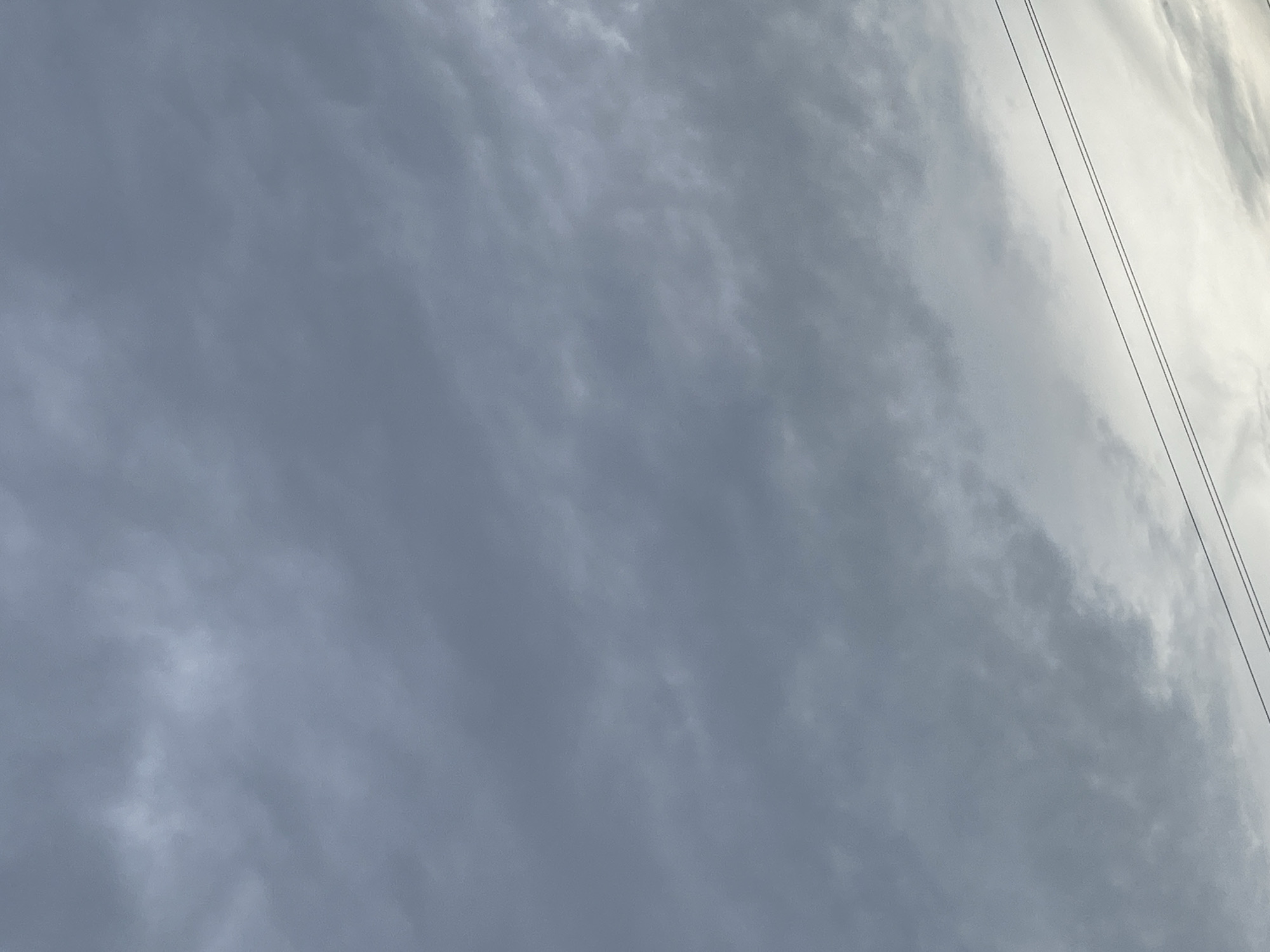 A low-angle shot of a cloudy sky is captured in Tinaquillo, Venezuela. The clouds are a mix of dark gray and lighter tones, indicating an overcast or potentially stormy day. The bottom of the frame shows two thin, dark power lines stretching horizontally across the image. The overall atmosphere is subdued and somewhat dramatic due to the heavy cloud cover.