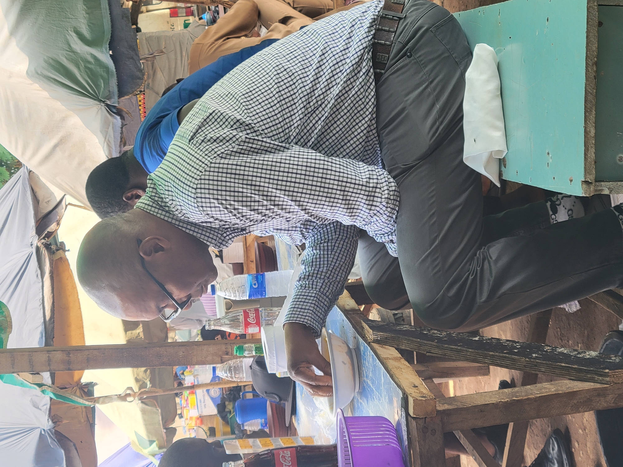 A man with glasses and a checkered shirt is leaning over a wooden table, eating from a white bowl. Several bottles of water and Coca-Cola are visible on the table. A second man wearing a blue shirt is partially visible in the background. The setting appears to be outdoors, possibly a roadside eatery in Abuja, Nigeria, as inferred from the visible local products and general ambiance. The scene appears to be a casual dining environment, with makeshift tables and overhead cover.