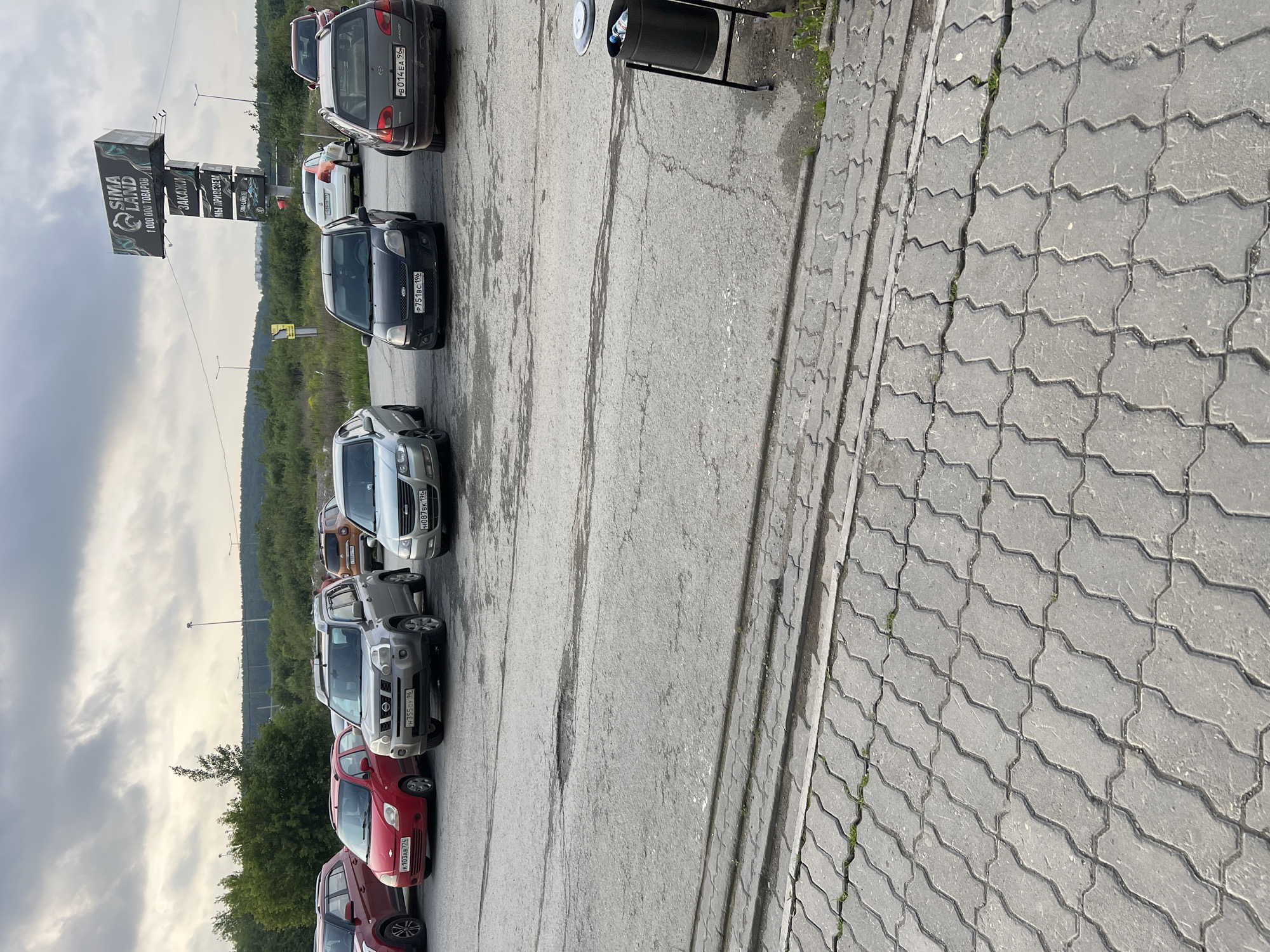 An eye-level outdoor medium shot captures a parking lot with several vehicles and a large billboard overhead under a cloudy sky. The parking lot surface is asphalt with visible cracks and patches, and a section of interlocking pavers is visible in the foreground. Cars parked in the lot include a red sedan, a silver SUV, a silver station wagon, a black hatchback, and a silver sedan. A black trash can stands on the right side of the frame. The billboard, which is mounted on a tall metal structure, displays advertisements for "SIMALAND" and "1 000 000 TOBAPOB". Trees and distant buildings are visible in the background.