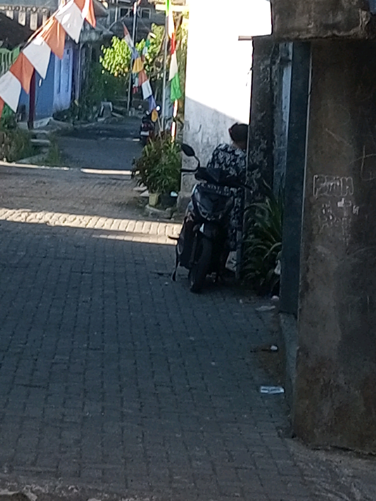 A person wearing a dark, patterned shirt is seated on a scooter parked near a building in Andongsili, Indonesia. The scooter is positioned on a paved street lined with brickwork. Banners in orange, white, and red are strung across the street, suggesting a local celebration or event. The background shows buildings and greenery, indicating a residential or public area. The overall scene is well-lit by natural sunlight.
