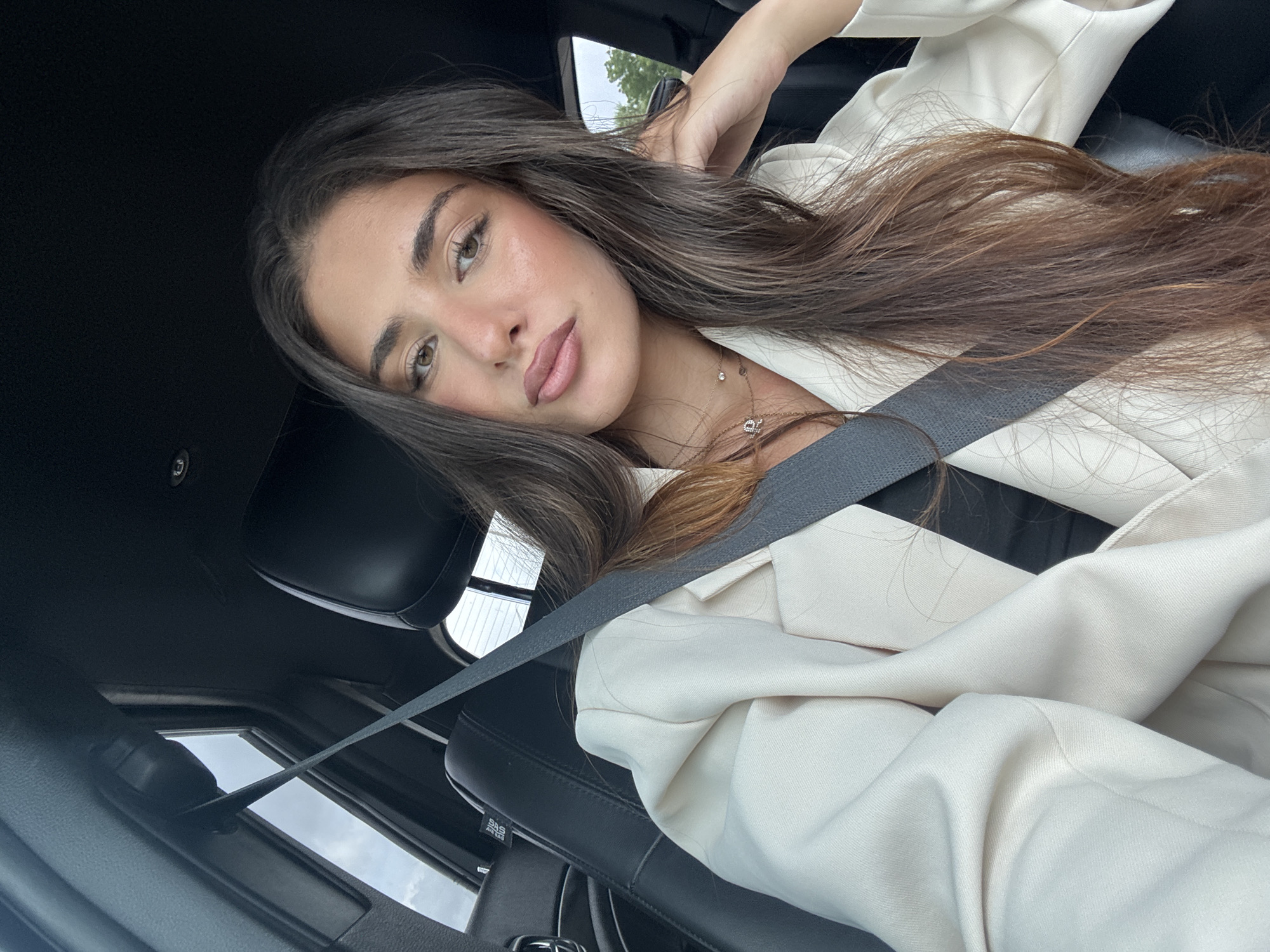 A young adult female is in the driver's seat of a car. She wears a beige blazer, a seatbelt, and a necklace. The location is presumed to be North Miami, Florida, United States.