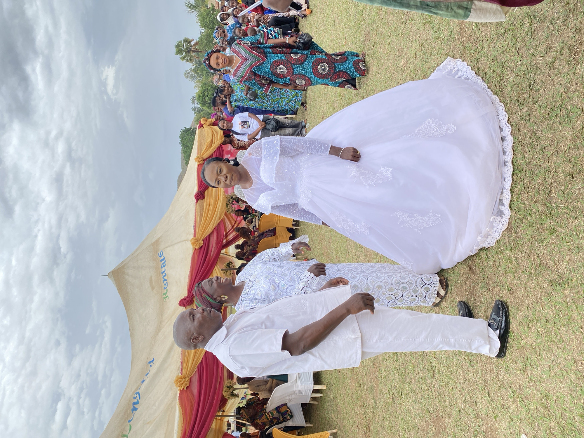 The image captures a wedding celebration held outdoors in Kuru Dungus, Nigeria. A bride in a white, long-sleeved gown with lace detailing stands alongside two people. An older woman in a patterned white dress and sandals is holding a glass. A bald man in a white suit and black shoes stands on the woman's right. They stand on a grassy area. Behind them is a large tent, decorated with yellow and red draping. People are sitting under the tent. A crowd of guests dressed in colorful clothing stands in the background. The sky is overcast.