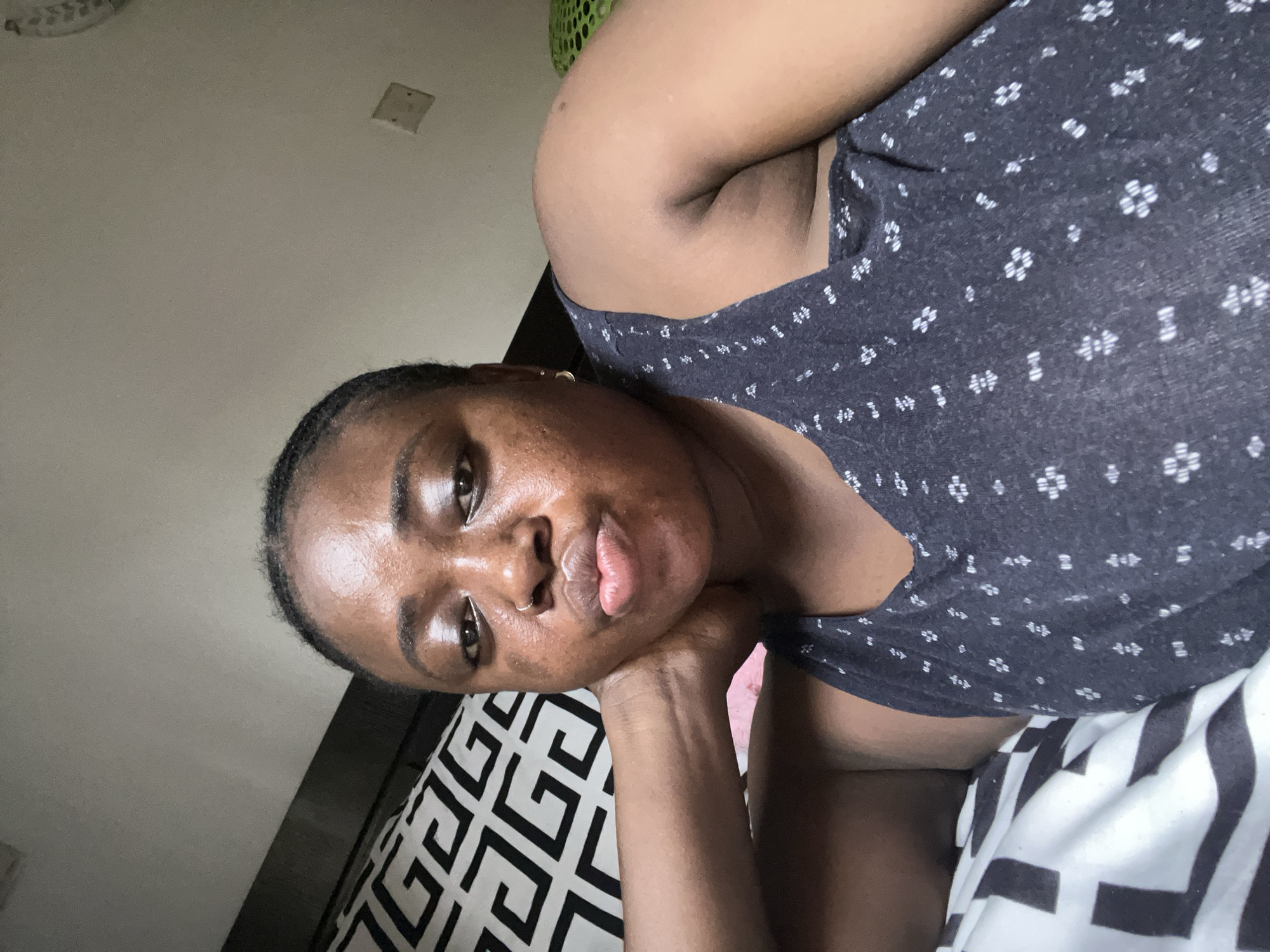 A brown-skinned woman with a dark gray shirt printed with small white flowers rests her face on her hand and gazes directly into the camera. Her dark hair is pulled back from her forehead. She is resting on a bed with a black and white patterned pillow. The background features a light beige wall and a visible light switch. There is no indication of the location in Abuja, Nigeria.