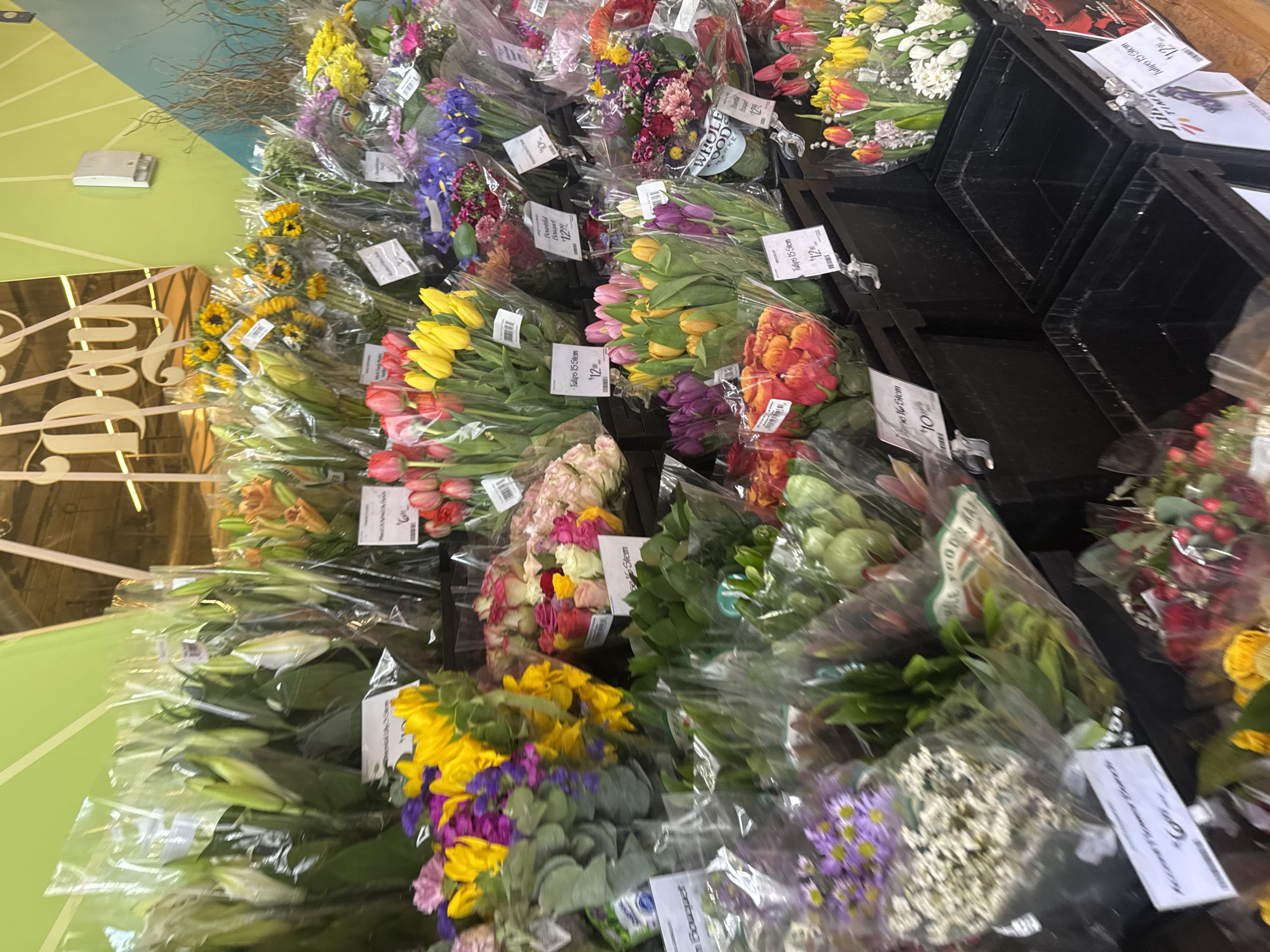 A variety of flowers, mostly bouquets, are displayed for sale on a table in what appears to be a retail setting, likely in Los Angeles, United States of America, given the OCR text 'WHOLE FOODS MARKET.' The flowers are arranged in clear plastic wraps and are accompanied by price tags. Many are labeled 'Tulips 15 Stem' with a price of '$12', while others are marked as 'Bouquet' at '$12.99.' There are also price tags indicating '10% off'. The flowers include tulips in various colors, sunflowers, lilies, and other mixed floral arrangements. They are organized in black containers on the table. The surrounding environment is somewhat blurred, but it appears to be a brightly lit store or market area.