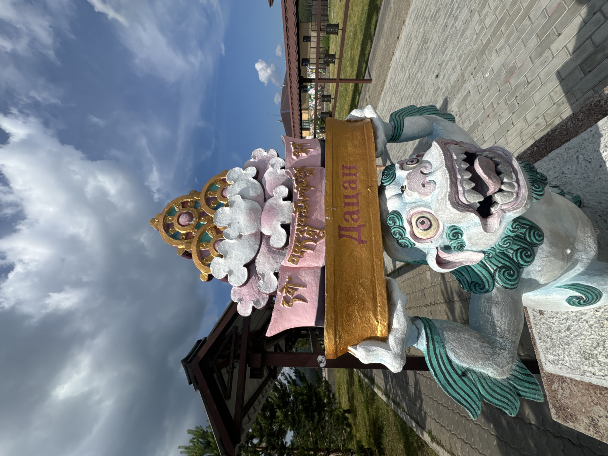 The image shows a brightly colored statue in an outdoor setting, likely a temple or monastery. The statue depicts a mythical creature, possibly a lion or dragon, supporting a decorative structure on its head. This structure features pink and white clouds, a golden banner with text in Cyrillic letters, and ornate golden and colorful details at the top. The creature itself is white with teal accents on its mane, wings, and tail, and has a gaping mouth with sharp teeth and a pink tongue.
The statue is positioned on a paved walkway, with grass and trees visible in the background. To the left, a wooden gazebo or shelter with a dark roof and brown posts is partially visible. Further back, a brown fence encloses a grassy area with what appear to be prayer wheels or decorative urns. Buildings with reddish-brown roofs are seen in the distance. The sky is blue with scattered white and dark clouds.
This statue is located at the Ivolginsky Datsan, a Buddhist monastery located in the Ivolginsky District of Buryatia, Russia, near Ulan-Ude. The inscription on the golden banner reads "Дацан," which means "Datsan" in Russian, referring to a Buddhist monastery.