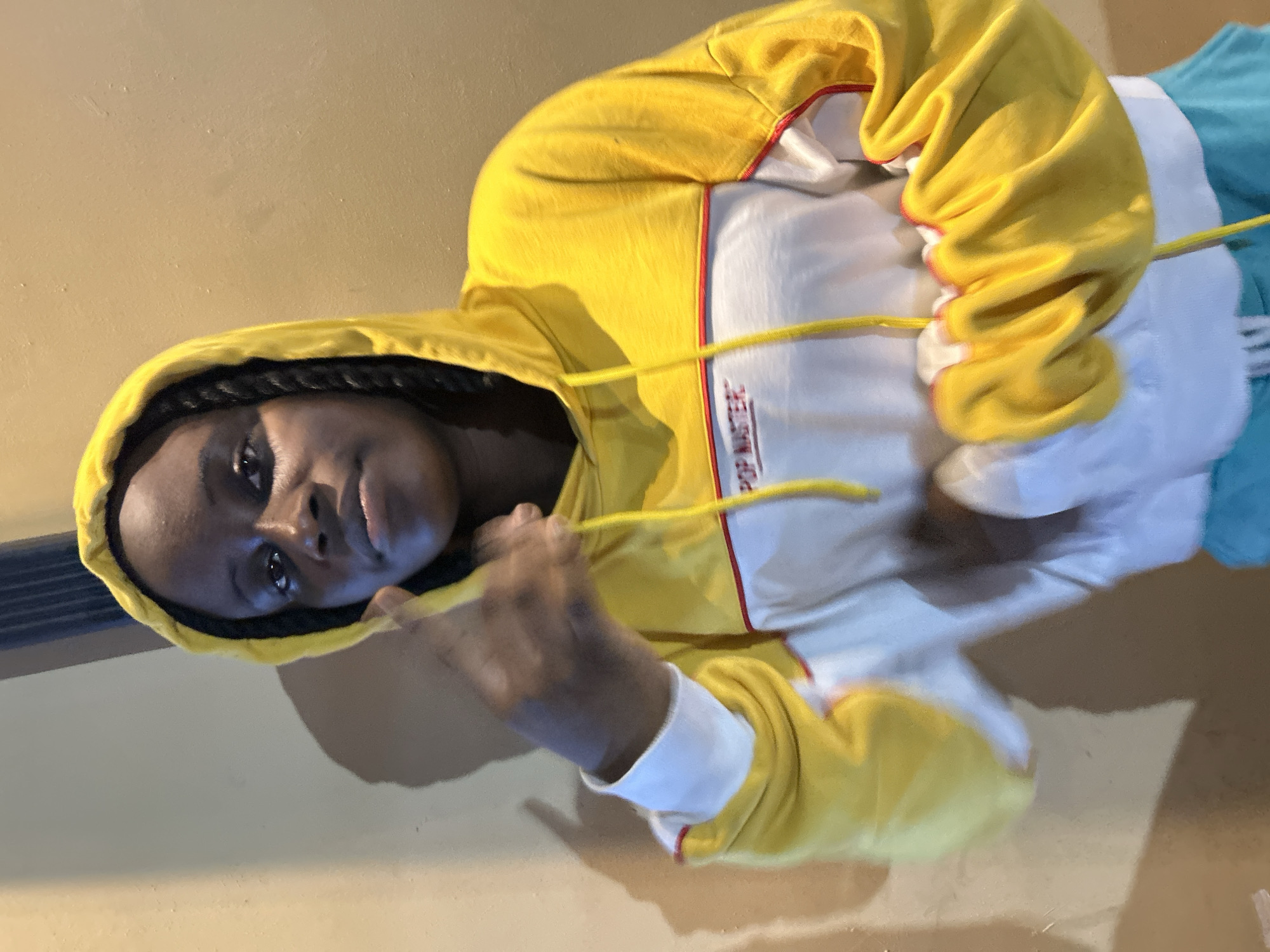 In the city of Ihiagwa, Nigeria, a young Black person, likely a teenager, is captured in a medium shot, standing against a plain, light brown wall. The individual is wearing a bright yellow hooded sweatshirt with white and red accents. The sweatshirt has the words "POP MASTER" embroidered in red on the front. Their hair is styled in dark braids that are visible from within the hood. The person's right hand is raised, with their index finger and thumb extended in a gesture that could be interpreted as a "shaka" or a similar hand sign. The lighting creates a shadow of the person on the wall behind them, adding depth to the scene. The overall impression is casual and relaxed, with the subject posing for the camera.