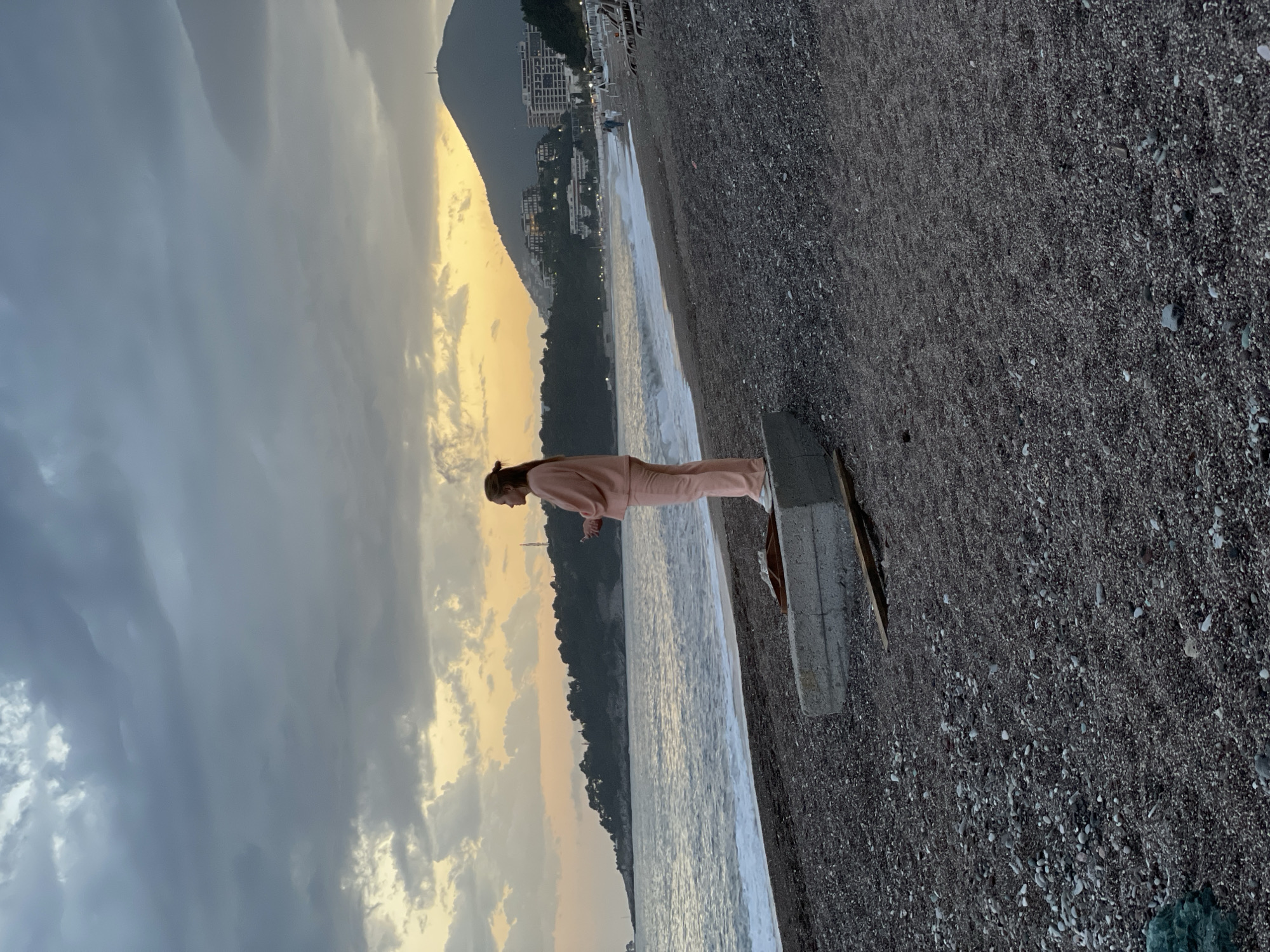 The photograph depicts a woman standing on a concrete pier-like structure, appearing to be at a beach in Rafailovići, Montenegro. The woman is wearing a pink tracksuit and is looking down toward the water. The beach is composed of dark pebbles, and the sea is calm with small waves. In the background, a mountain range meets the coastline, and some buildings are visible. The sky is overcast with a combination of grey clouds and a golden hue near the horizon, indicating either sunrise or sunset.