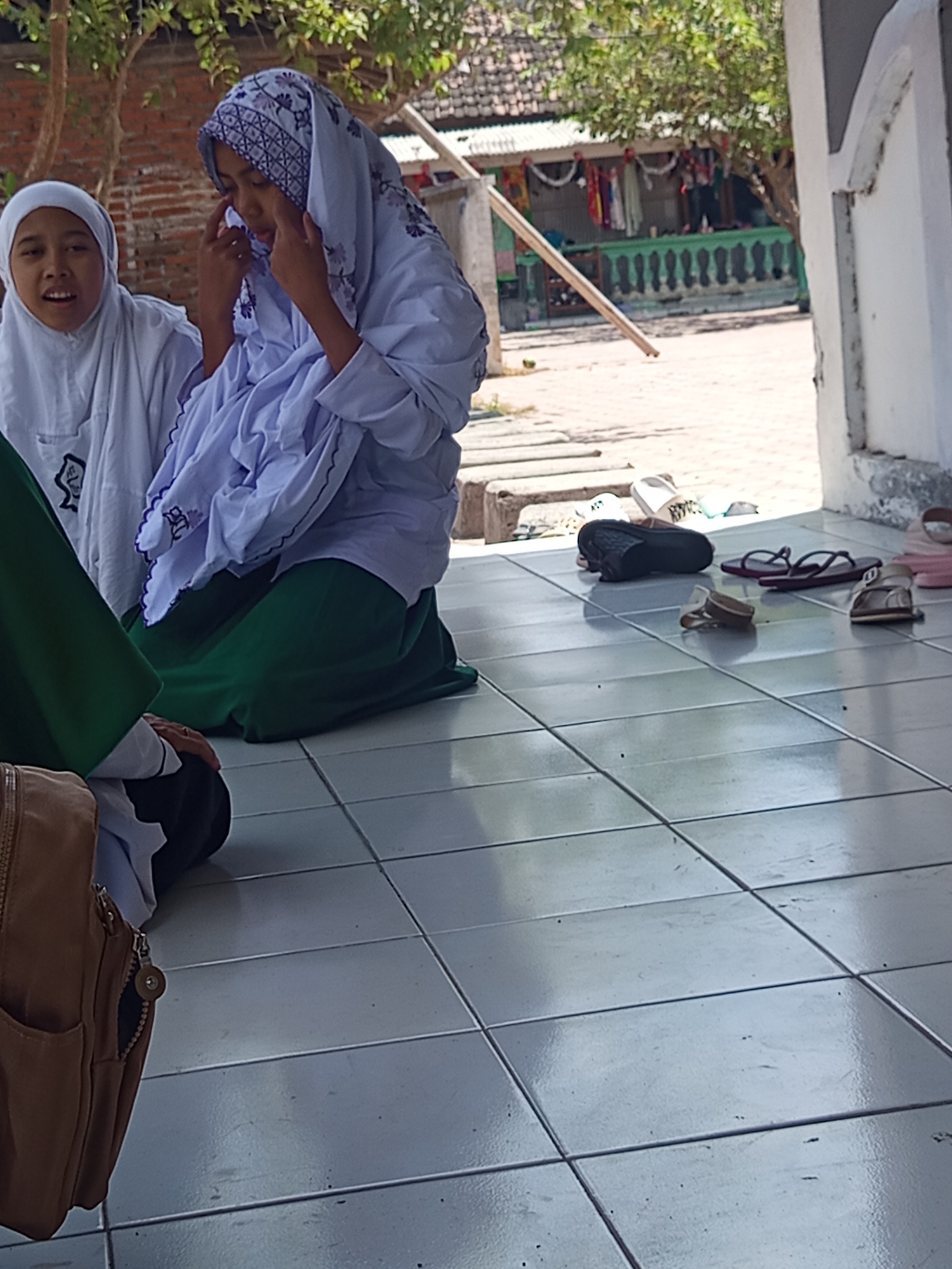 This is a medium shot, eye-level, vertical photograph taken outdoors in Lumutan, Indonesia. The foreground features a tiled floor. On the floor, several pairs of women's footwear are scattered, including a pair of black flip-flops, a pair of maroon flip-flops, and a pair of beige and brown sandals. In the middle ground, two young females are kneeling on the tiled area. The female on the left wears a white hijab and a white garment. The female on the right wears a white hijab with a purple floral pattern and a purple and white patterned garment over a long green skirt. She is touching her face with her right hand, her eyes are closed, and her head is tilted down, suggesting she may be praying or distressed. Behind the kneeling individuals, in the background, a tree with green foliage is visible. Further back, a building with a green railing and what appears to be clothes hanging on a line is present. The scene is illuminated by natural sunlight, casting shadows on the tiled floor. A brown backpack rests on the floor in the lower left corner of the image.