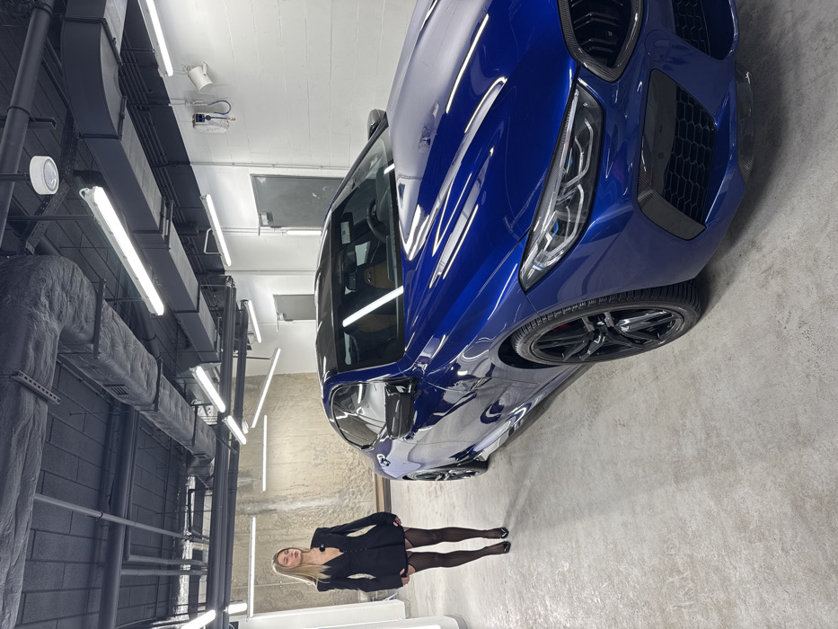 This image captures an indoor professional car detailing or showroom facility, identified as being in Warsaw, Poland. The environment features a light gray concrete floor and a black industrial ceiling from which exposed ductwork, pipes, and multiple linear fluorescent light fixtures are suspended. The left wall exhibits a light-colored, textured concrete finish, contrasting with the white-painted paneling on the right wall, which includes two gray metal doors and a wall-mounted utility unit with coiled hoses.
A highly polished, dark metallic blue BMW M8 two-door sports coupe is prominently positioned in the mid-to-right foreground, angled with its front toward the viewer. The vehicle features black multi-spoke wheels, visible red brake calipers, and elements of carbon fiber on its exterior, including the roof and front bumper trim. The interior, visible through the windshield, shows light brown leather upholstery.
To the left of the vehicle, a blonde female subject stands with her body slightly angled towards the left, her gaze directed off-camera. She is dressed in a black, short-length blazer-style garment with prominent shoulder pads, sheer black stockings, and black pointed-toe high heels. A small device, consistent with a lapel microphone, is affixed to her left lapel.
