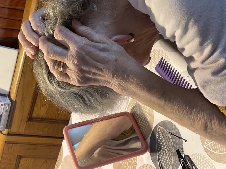 An overhead shot captures an older person, likely female, engaged in hair care within an indoor setting. The individual's head is bent forward, revealing grey hair, which is being manipulated by both hands. A gold-colored ring is visible on the ring finger of the left hand. The person is wearing a light-colored, long-sleeved top.
The scene takes place on a surface covered by a patterned placemat featuring abstract leaf-like shapes. Scattered on this surface are a purple comb, a small pink-framed mirror reflecting an arm, and several black hairpins or clips. In the background, wooden cabinets and a partial white object with illegible text are visible. The lighting suggests an indoor environment, but the specific time of day is not discernible. The activity appears to be a routine personal grooming task. No visible text can be clearly read. The location context provided for this scene is Dosso (Ciappanico), Italy.