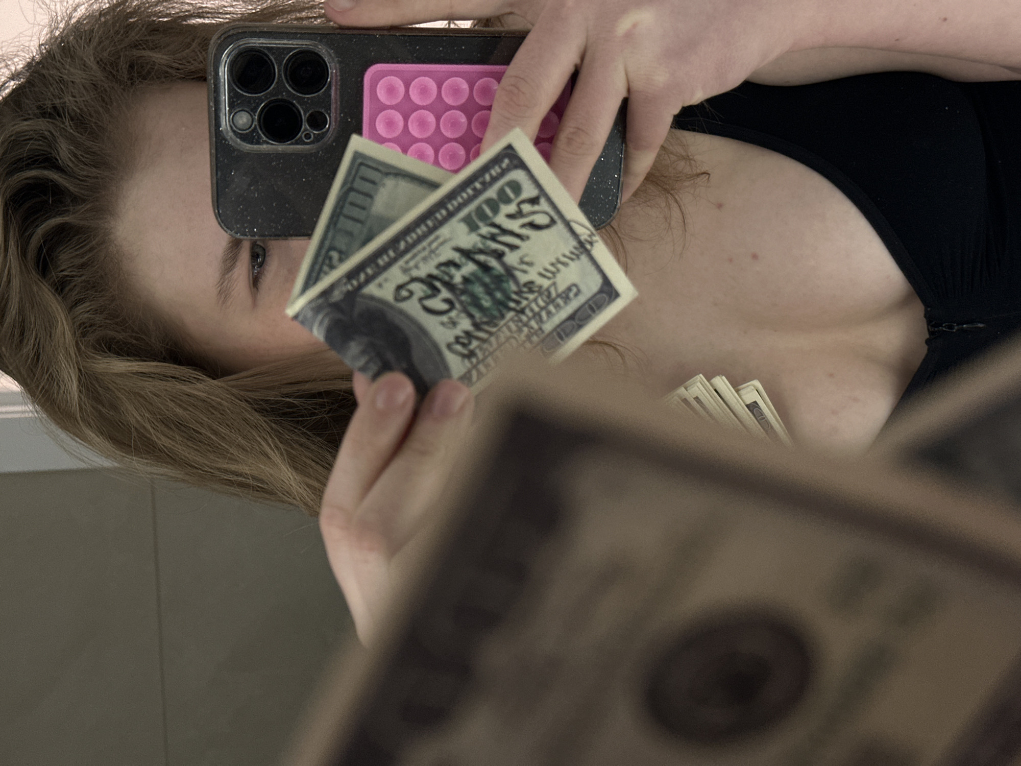 A person is holding a smartphone with a black case and a pink pop-it fidget toy attached to it, obscuring their face, while also holding US hundred-dollar bills in front of the camera. A stack of US hundred-dollar bills is visible in the person's cleavage. The background appears to be a reflection in a mirror, possibly in a bathroom or similar interior space. The location in Kazan, Russia cannot be ascertained from the image.