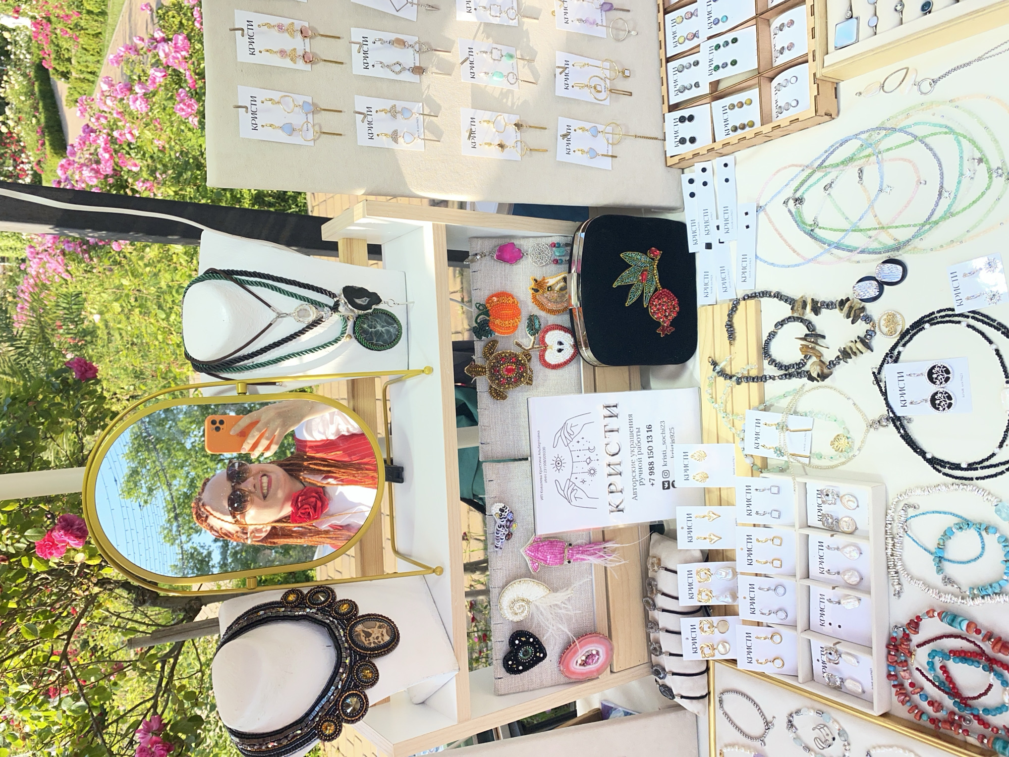 This image depicts an outdoor market stall in Sochi, Russia, with a focus on handmade jewelry. A woman with braided red hair and sunglasses is taking a selfie in a gold-framed mirror, holding an orange smartphone. She is wearing a red vest over a white shirt and a red flower accessory around her neck. The stall displays a variety of necklaces, earrings, bracelets, and brooches. Some of the jewelry pieces are presented on white mannequins, while others are arranged in display cases and on hanging cards. The background reveals a lush green garden with blooming pink roses, characteristic of the natural beauty found in Sochi. The overall scene suggests a vibrant and artisanal atmosphere at a local market.
