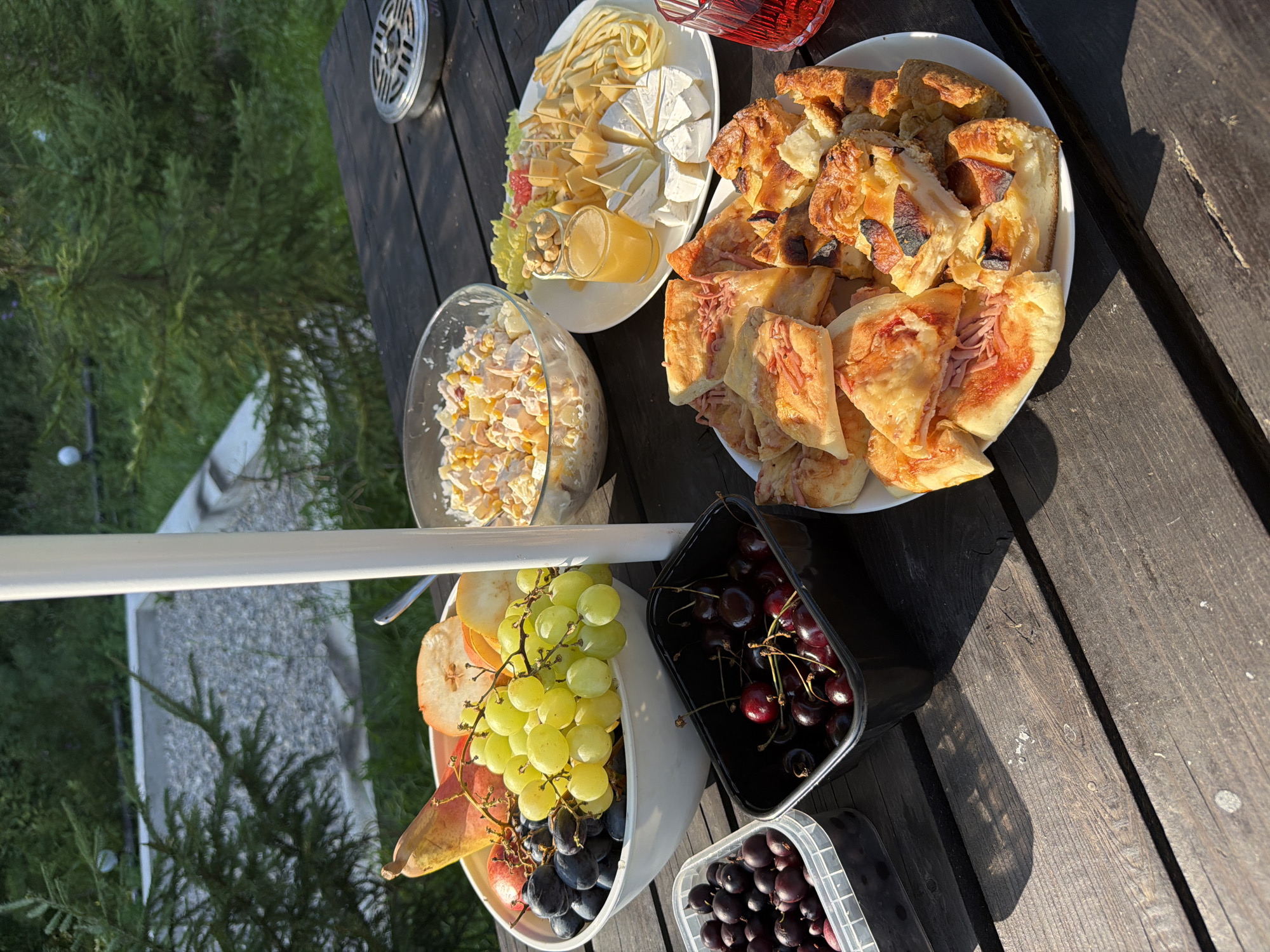 This is an overhead, slightly angled medium shot of a picnic spread on a dark wooden table outdoors, likely in Kuliki, Russia. The table is laden with various food items and a white pole, possibly part of an umbrella or tent, bisects the image vertically.
In the foreground to the left are two plastic containers. The container on the left holds dark berries, possibly blueberries or blackcurrants. The container to the right holds dark red cherries.
To the right of the containers is a large white bowl overflowing with a variety of fruits, including green and dark grapes, sliced pears, and red apples.
Behind the fruit bowl is a glass bowl filled with a creamy salad containing corn and other ingredients.
To the right of the salad is a white plate with an assortment of cheese, including wedges of brie and cubes of yellow cheese, accompanied by what appears to be small nuts in a shot glass of orange juice. There are also some pieces of what looks like grapefruit or pomelo on the plate, garnished with small toothpicks.
Further to the right, and closer to the foreground, is a white plate piled high with sliced baked goods. These appear to be a mix of savory items, including what looks like small pizzas with cheese and ham, and sweet items, such as apple pie or tart slices with browned crust.
A glass of red liquid, likely juice or wine, is visible on the far right edge of the table.
The background shows lush green trees and some structures, suggesting a natural setting. The lighting indicates it is daytime, with sunlight casting shadows on the wooden table. There are no people visible in the image.