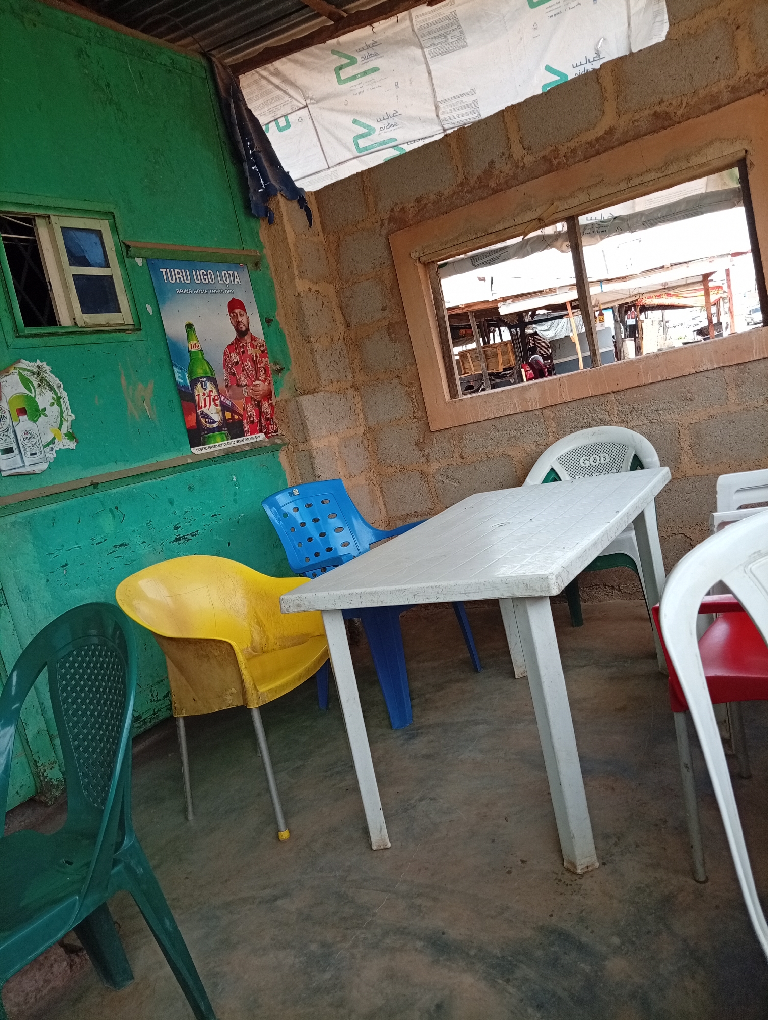 The image depicts an interior scene, likely in a public setting within Jos, Nigeria. The walls are a mix of painted green wood and exposed concrete blocks. A small window is visible on the green wall, and a larger window frames a view of a street with market stalls. A plastic table with white legs sits centrally, surrounded by mismatched plastic chairs in blue, yellow, green, white and red. A "Life" beer poster is affixed to the green wall, next to an "orijin" poster. The floor is a mottled grey. The setting appears to be a low-cost eatery or bar, possibly in a commercial district.